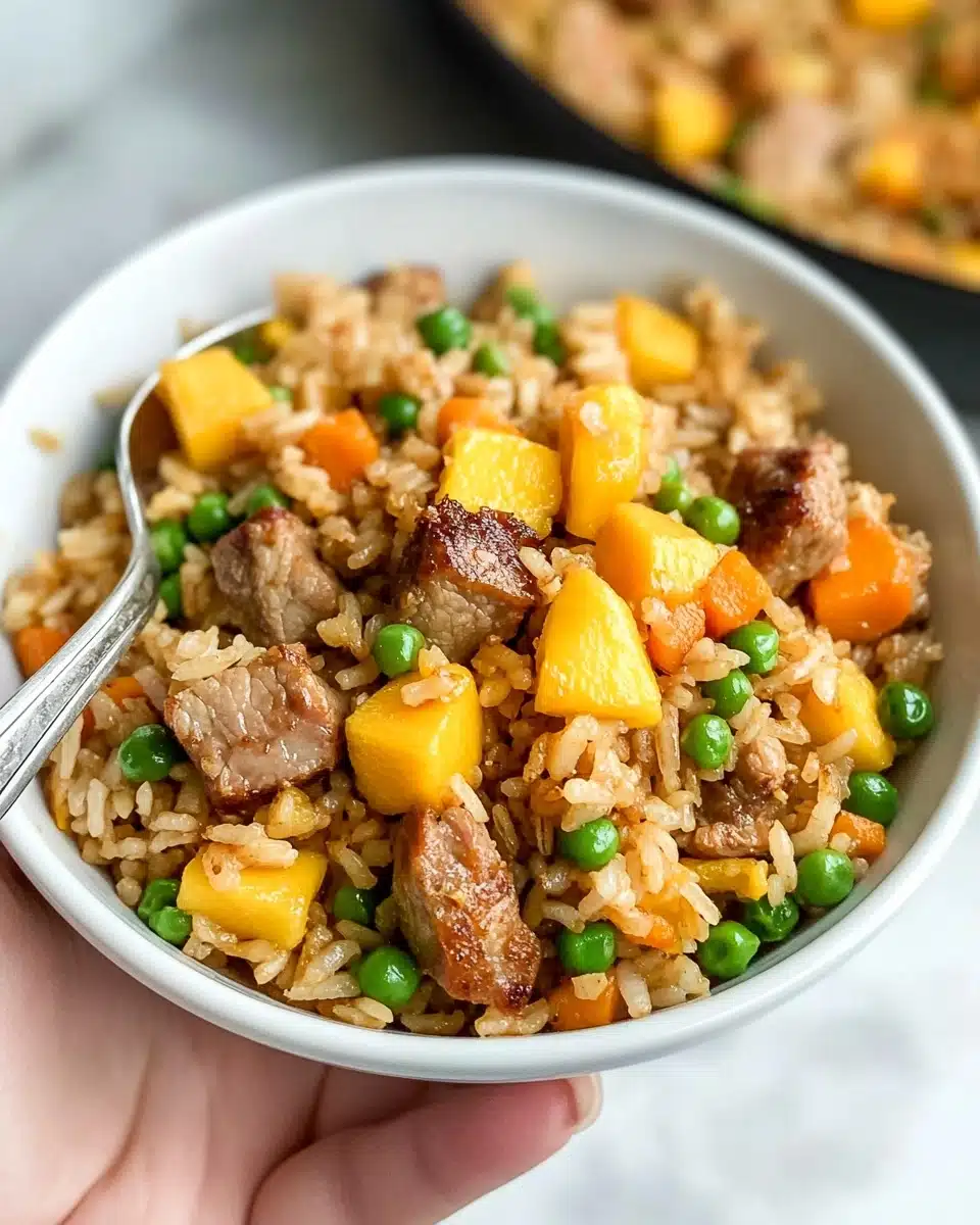 A white bowl is filled with fried rice mixed with small green peas, diced orange carrots, and bright yellow mango chunks. On top, there are several pieces of browned cooked meat with a slightly crispy texture. A silver spoon is placed inside the bowl on the left side. The bowl is held by a woman's hand over a white marbled surface, and a blurred pan of more cooked rice with vegetables is visible in the background. photo taken with an iphone --ar 4:5 --v 6.1