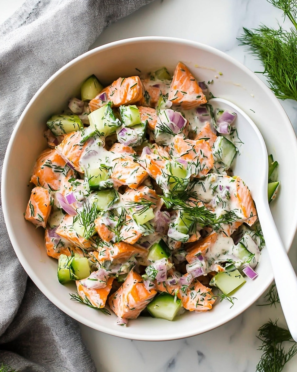 A white bowl holds a mix of cooked salmon pieces in light pink shades with slight browned edges, mixed with small chopped green celery, pale green cucumber, and finely diced red onion. The dish is sprinkled with finely chopped fresh dill herbs adding green flecks all over. A creamy white sauce with small herb bits is seen lightly coating the ingredients, creating a moist texture. A large white spoon rests inside the bowl on the right side. Around the bowl, there is a white marbled surface with a small glass bowl of extra sauce to the right and some fresh dill sprigs on the upper right. A gray textured cloth is partly seen on the left. Photo taken with an iphone --ar 4:5 --v 6.1