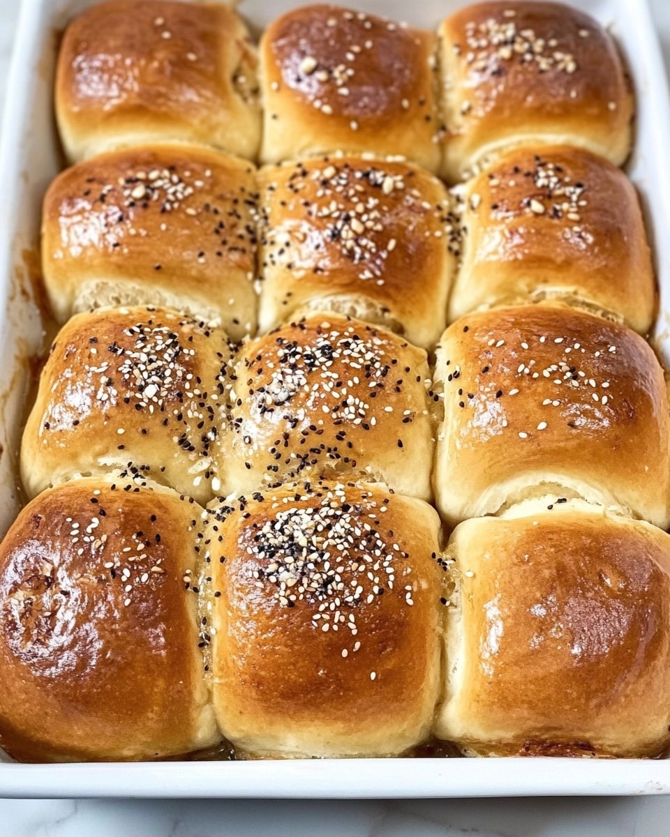 A white square baking dish holds twelve soft, golden brown rolls arranged in a 3 by 4 grid. Each roll has a slightly wrinkled, shiny top sprinkled evenly with a mix of white sesame seeds, black seeds, and small bits of dried garlic and salt. The rolls are touching each other with soft, slightly puffed sides. The dish sits on a white marbled surface. Photo taken with an iphone --ar 4:5 --v 6.1