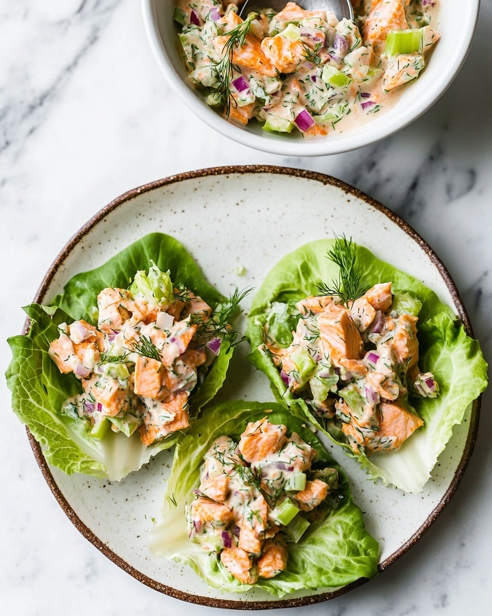 The image shows three lettuce wraps placed on a white plate with a brown rim, resting on a white marbled surface. Each wrap has a bright green leaf base, topped with a creamy mixture of light orange cooked salmon pieces mixed with small chopped pieces of green celery and red onion. Above the plate is a white bowl filled with more of this creamy salmon mixture, with a silver spoon resting inside it. The colors are fresh and vibrant, with the pink-orange salmon, green celery, red onion, and dill bits standing out clearly. Photo taken with an iphone --ar 4:5 --v 6.1