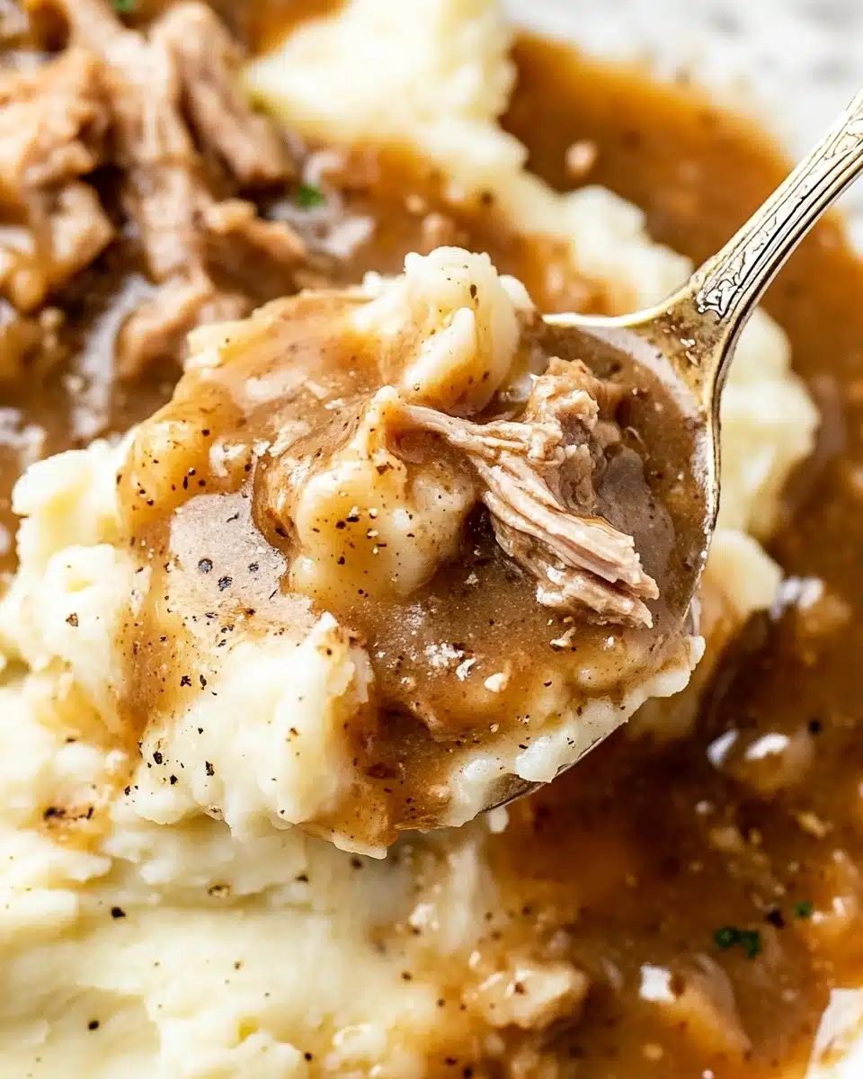 A close-up view of a spoon lifting creamy mashed potatoes topped with thick brown gravy and tender pieces of shredded meat. The mashed potatoes are soft and smooth, pale white in color, sitting beneath a rich, glossy brown sauce with visible black pepper specks. The shredded meat is cooked, light brown, and mixed into the gravy, creating a layered texture with the mashed potatoes. The background is a white marbled surface with more of the mashed potatoes, gravy, and meat blurred softly behind the spoon. photo taken with an iphone --ar 4:5 --v 6.1