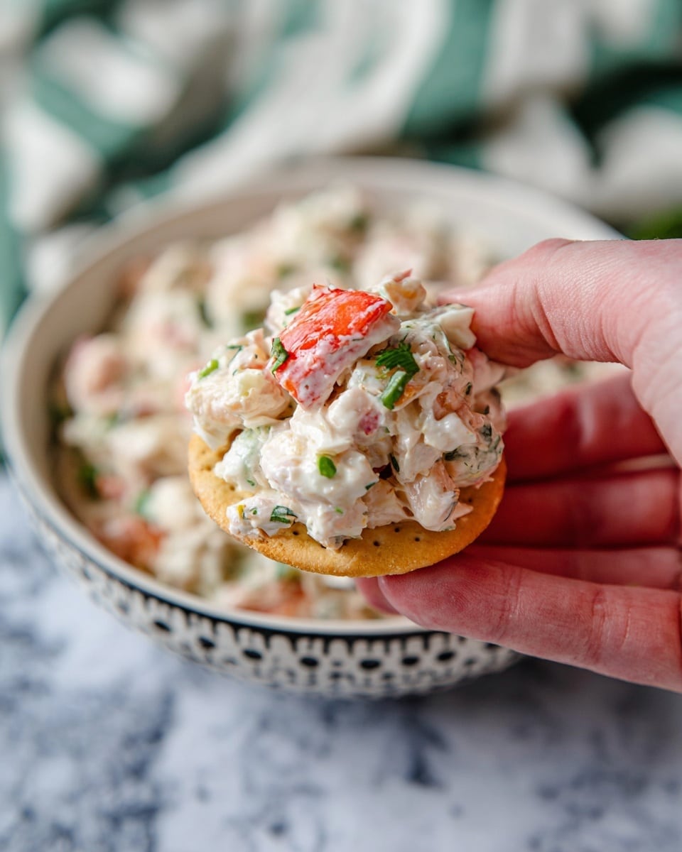 A close-up view shows a woman's hand holding a round, crispy golden chip topped with a creamy white salad that has pieces of red crab meat, small shrimp, and finely chopped green herbs. In the background, there is a white bowl filled with the same creamy salad with visible pieces of crab meat and herbs, resting on a white marbled surface with a blurred green-striped cloth behind it. The colors include the golden chip, white creamy salad with red and pale pink seafood, and green herbs. photo taken with an iphone --ar 4:5 --v 7