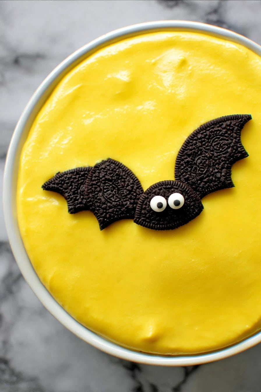 No Bake Boston Cream Halloween Pie, Halloween dessert, no bake pie recipe, easy Halloween treats, festive pumpkin pie - A round dessert in a white ceramic dish has a smooth, bright yellow top layer that looks creamy and shiny. On this yellow surface are two small decorations shaped like bats, made with halves of dark chocolate sandwich cookies for wings and two small white candy eyes on a chocolate base forming the face. The dish sits on a dark cloth background. photo taken with an iphone --ar 2:3 --v 7