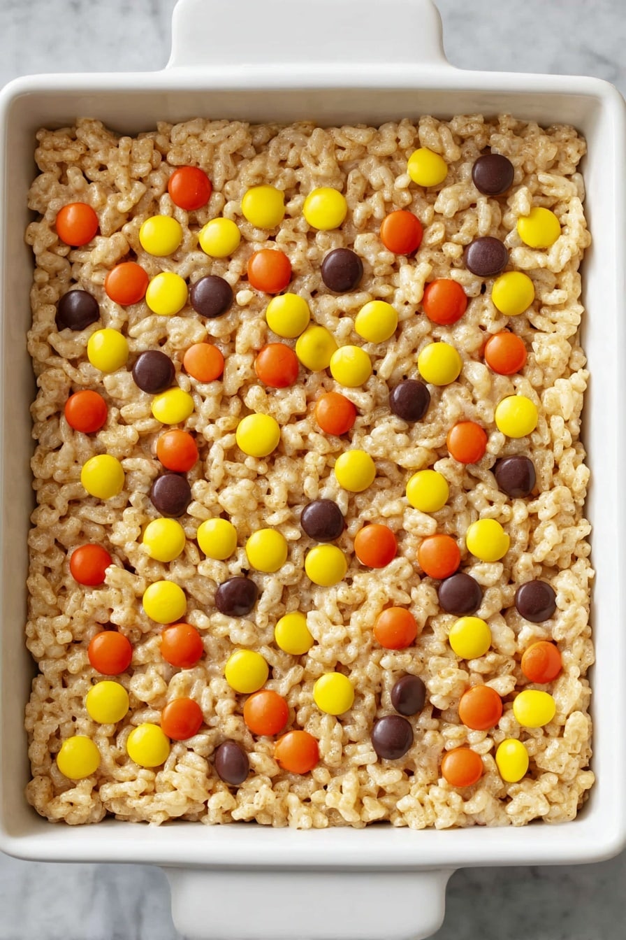 Chocolate Peanut Butter Rice Krispie Treats, peanut butter and chocolate dessert, homemade Rice Krispie treats, easy no-bake treats, kid-friendly snack - A single thick layer of light golden crispy puffed rice cereal mixed with melted marshmallow is pressed into a white square dish, decorated on top with scattered small round candies in bright orange, yellow, and dark brown colors. The texture of the cereal layer looks soft and sticky but holds together, and the candies are evenly spread out on the surface against a white marbled background. Photo taken with an iphone --ar 2:3 --v 7