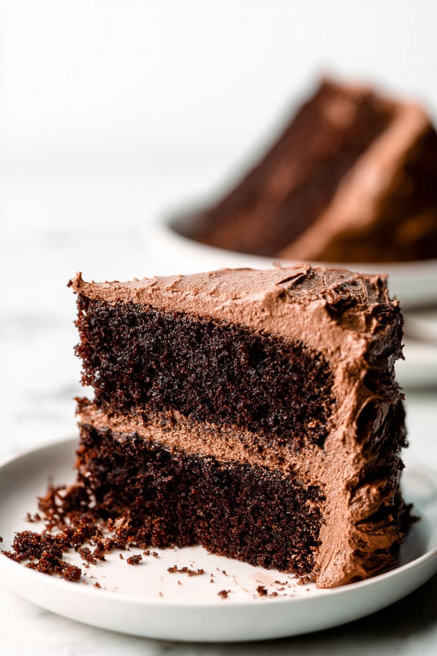 Gluten Free Vegan Chocolate Cake, vegan cake recipes, gluten free dessert, dairy free chocolate cake, plant-based chocolate cake - A close-up view of a two-layer chocolate cake slice on a white plate, placed on a white marbled surface. The cake layers are dark brown and moist with a rough texture. Between the layers is a medium brown chocolate frosting that looks creamy and smooth. The outside of the cake slice is covered in the same chocolate frosting, which is thick and slightly swirled, with rough edges. Some frosting has spilled onto the plate. In the background, there is a blurred image of another chocolate cake slice on a white plate. Photo taken with an iphone --ar 2:3 --v 7
