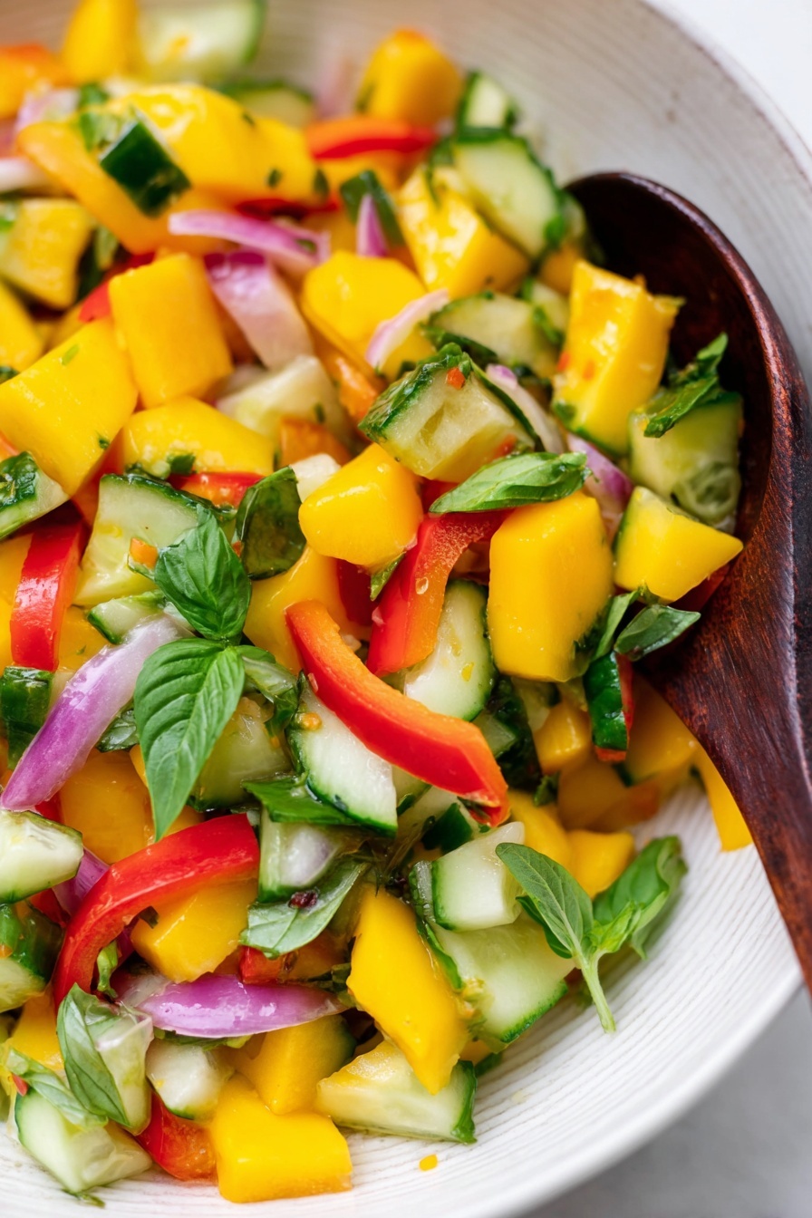 Mango Salad with Fresh Basil and Lemon Ginger Dressing, fresh mango salad recipe, healthy mango salad, citrus basil fruit salad, quick tropical fruit salad - A close-up of a colorful salad in a white bowl with a white marbled background, showing bright yellow mango cubes, green cucumber pieces, red bell pepper chunks, thin slices of light purple onion, and fresh green basil leaves mixed together. A dark wooden spoon is resting inside the bowl, partly covered by the salad. The colors are vibrant and fresh, with the textures of the juicy mango, crunchy cucumber, and leafy greens clearly visible. Photo taken with an iphone --ar 2:3 --v 7