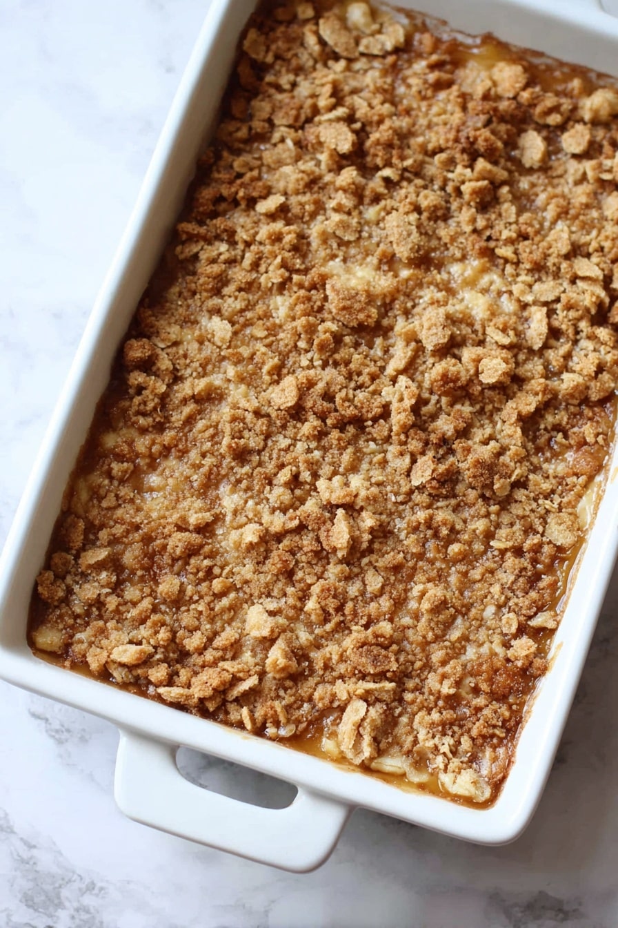 Pumpkin Dump Cake, easy pumpkin dessert, fall dessert recipes, quick pumpkin cake, pumpkin dessert ideas - The image shows a baked dessert in a white rectangular dish with two handles. The dessert has one main layer with a golden brown crumbly topping made of crushed pieces with a rough, uneven texture. The color is mixed shades of light to medium brown, giving a crunchy and toasted look. The dish is placed on a white marbled surface. Photo taken with an iphone --ar 2:3 --v 7