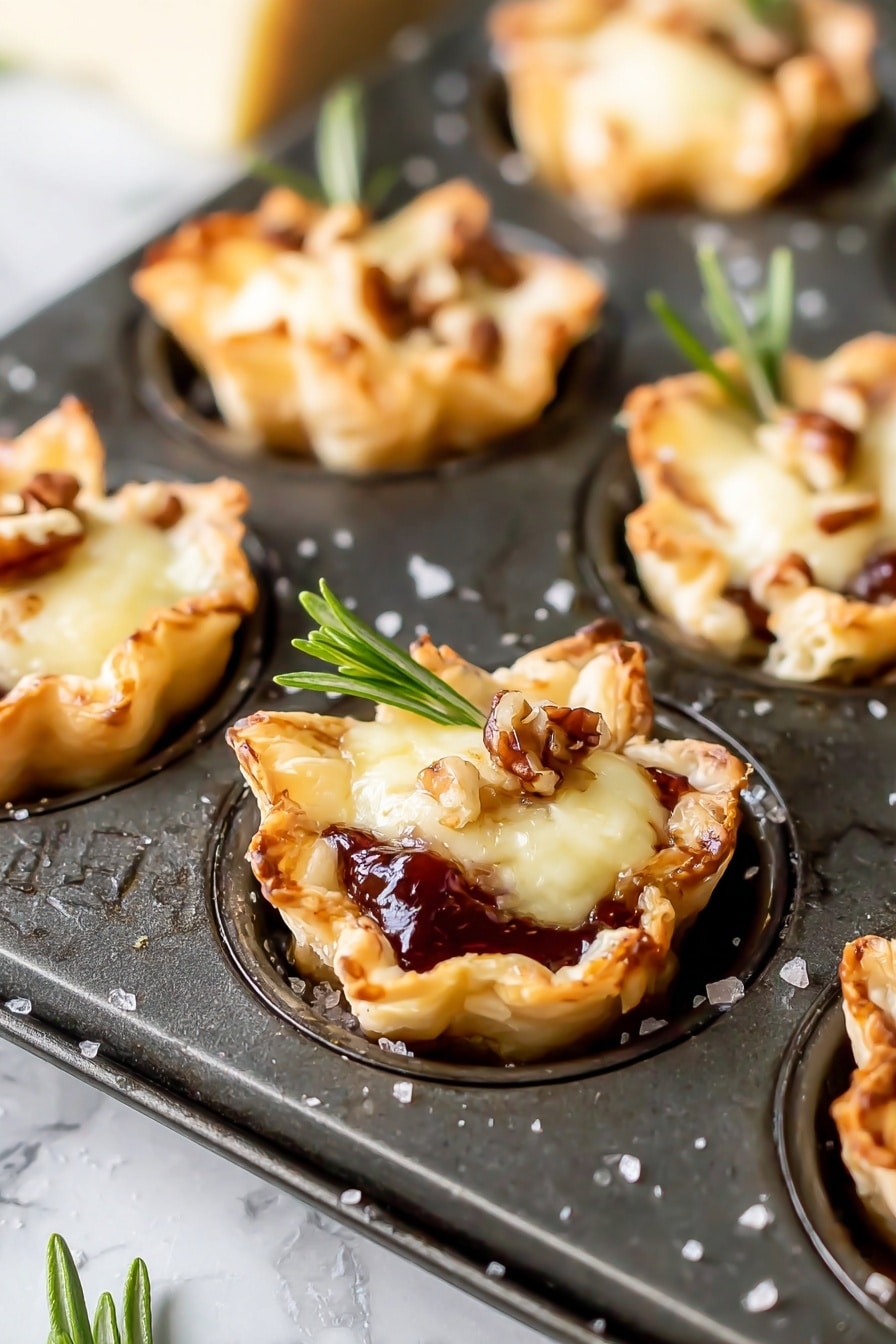 Cranberry Brie Bites, Cranberry Brie Appetizer, Puff Pastry Brie Bites, Holiday Party Snacks, Easy Cranberry Brie Bites - The image shows mini tartlets baked in a dark metal muffin tray, each tartlet having three main layers. The bottom layer is a golden-brown, flaky pastry shaped into small cups with slightly crispy edges. The middle layer is a glossy, dark red jam or fruit preserve that peeks out from beneath the top. The top layer is a melted, creamy pale yellow cheese, topped with small brown nut pieces and a fresh green rosemary sprig for garnish. The background is a white marbled texture, and some salt granules are scattered around the tray. photo taken with an iphone --ar 2:3 --v 7