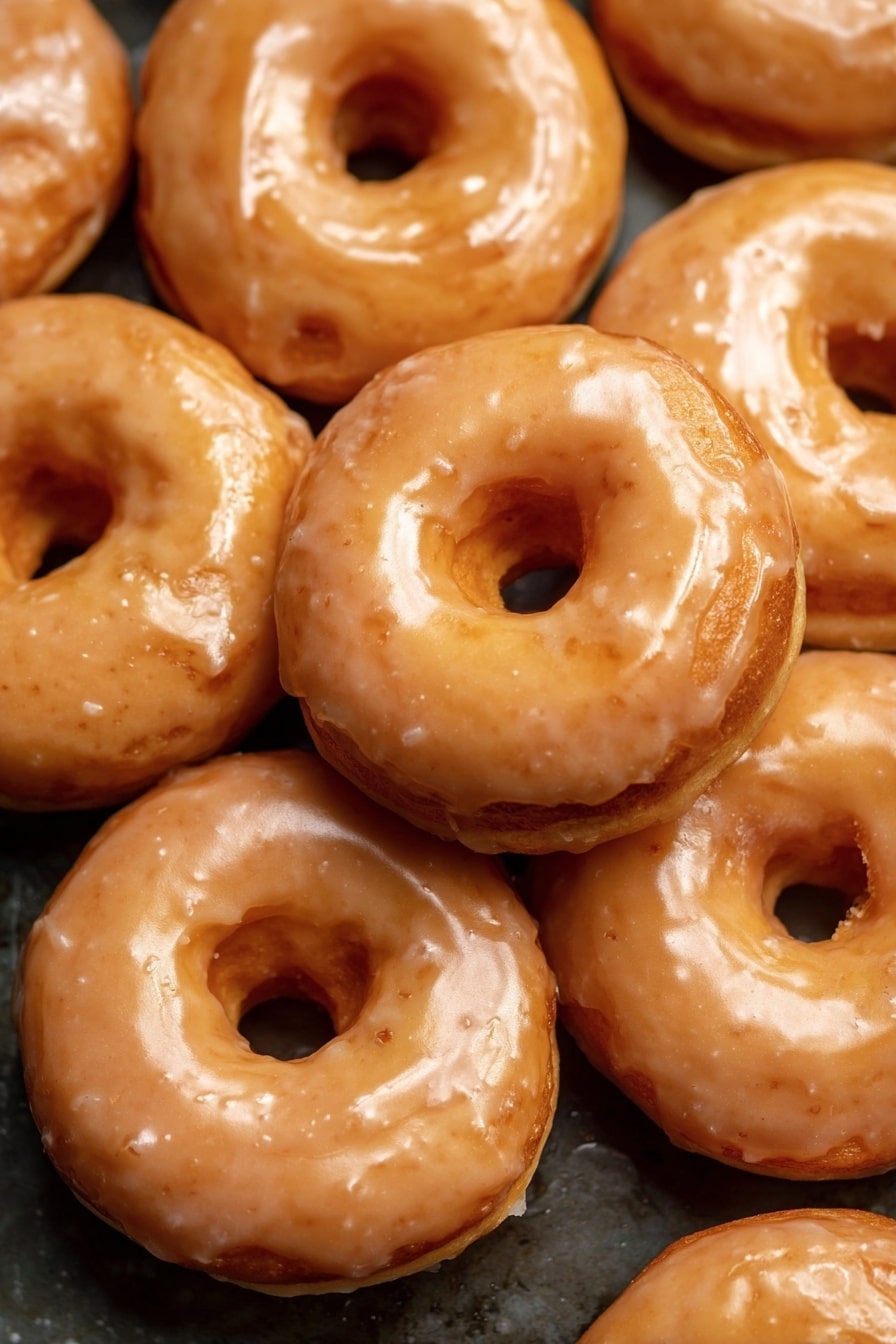 Vegan Pumpkin Donuts with Cinnamon Maple Glaze, Vegan pumpkin donuts, healthy pumpkin donut recipe, fall vegan desserts, baked pumpkin donuts - The image shows a close-up of many round donuts stacked together. Each donut has two visible layers: a base layer that is a golden brown color with a soft texture and a shiny glaze layer on top that is light tan and slightly translucent. The glaze evenly covers the top and sides of each donut, giving them a glossy look. The donuts have a round hole in the middle, and their surface shows slight bumps and smooth spots. The background is a dark surface contrasting with the shiny donuts. photo taken with an iphone --ar 2:3 --v 7