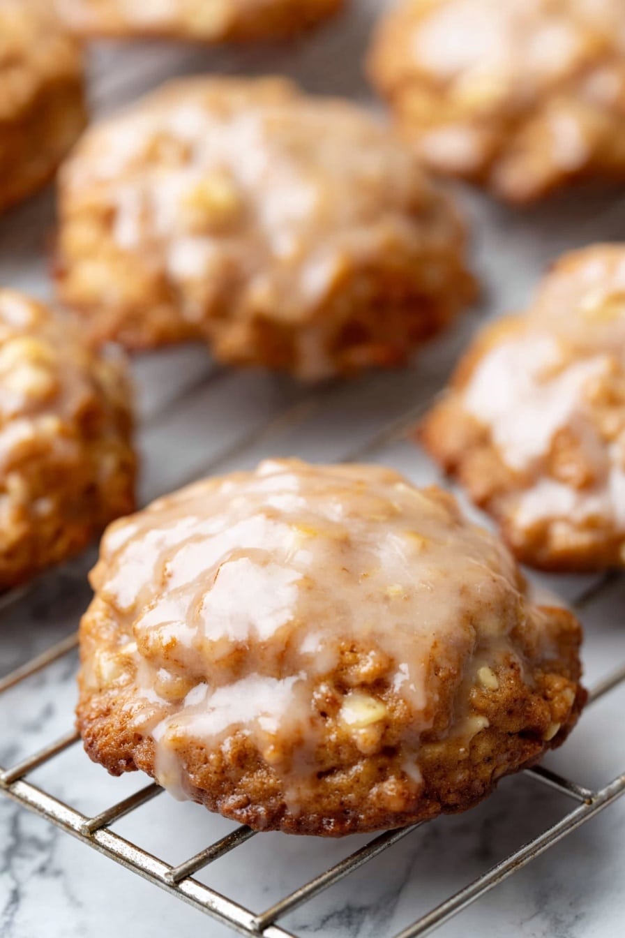 Apple Fritters, Apple Fritters Recipe, Easy Apple Fritters, Crispy Apple Fritters, Homemade Apple Fritters - The image shows several round cookies on a cooling rack. Each cookie has a shiny, light brown glaze on top with a slightly uneven texture. The cookies have a golden brown color, with a rough surface and visible small pieces inside, likely nuts or oats. The cooling rack underneath is metal and placed over a white marbled surface. The cookies look soft and have a slightly wavy edge. Photo taken with an iphone --ar 2:3 --v 7