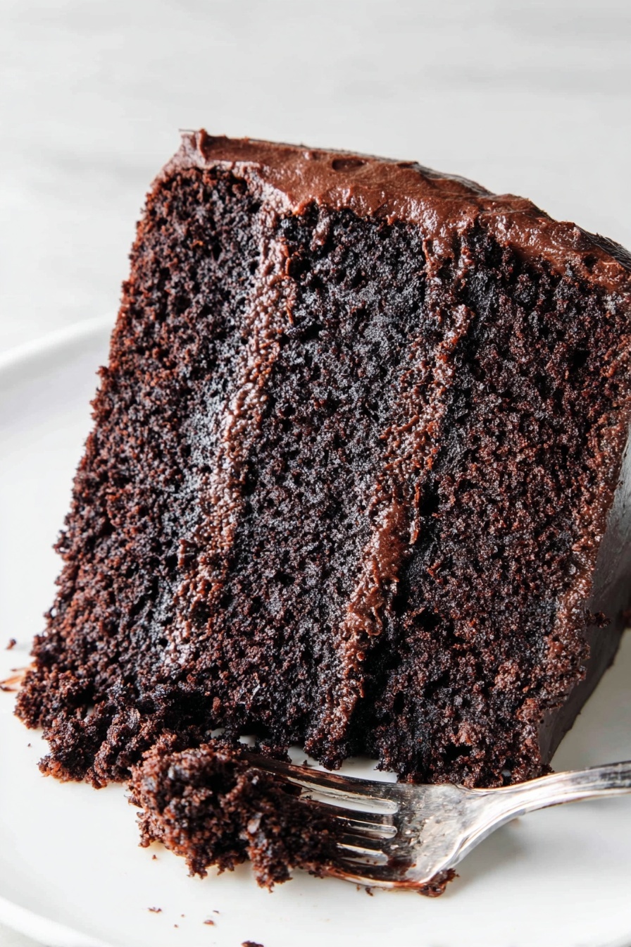 The Most Amazing Chocolate Cake, best chocolate cake, moist chocolate cake, chocolate cake recipe from scratch, rich chocolate cake - The image shows a slice of chocolate cake on a white plate placed on a white marbled surface. The cake has two thick layers of moist, dark brown chocolate sponge with a rich, glossy chocolate frosting layer in the middle. The outside of the cake is covered with a smooth, shiny chocolate frosting. A silver fork with some chocolate frosting rests on the plate next to the slice. Photo taken with an iphone --ar 2:3 --v 7