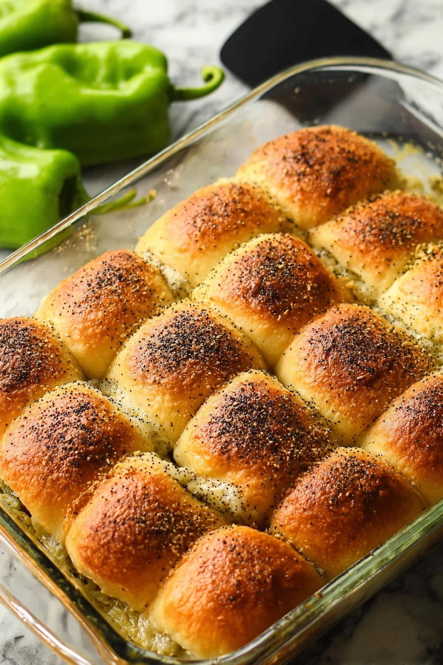 Santa Fe Turkey Sliders, Turkey Sliders with Green Chiles, Spicy Turkey Slider Bites, Easy Turkey Slider Appetizer, Game Day Turkey Sliders - The image shows a clear glass baking dish filled with twelve golden brown sandwich rolls arranged in three rows of four. Each roll has a slightly crispy top with visible poppy seeds sprinkled over them, giving a textured look. The edges of the rolls touch each other, and a melted, creamy layer is seen bubbling around the sides beneath the rolls. In the background, there are some green fresh peppers resting on a white marbled surface. A black spatula is placed beside the dish. The photo is taken with an iphone --ar 2:3 --v 7