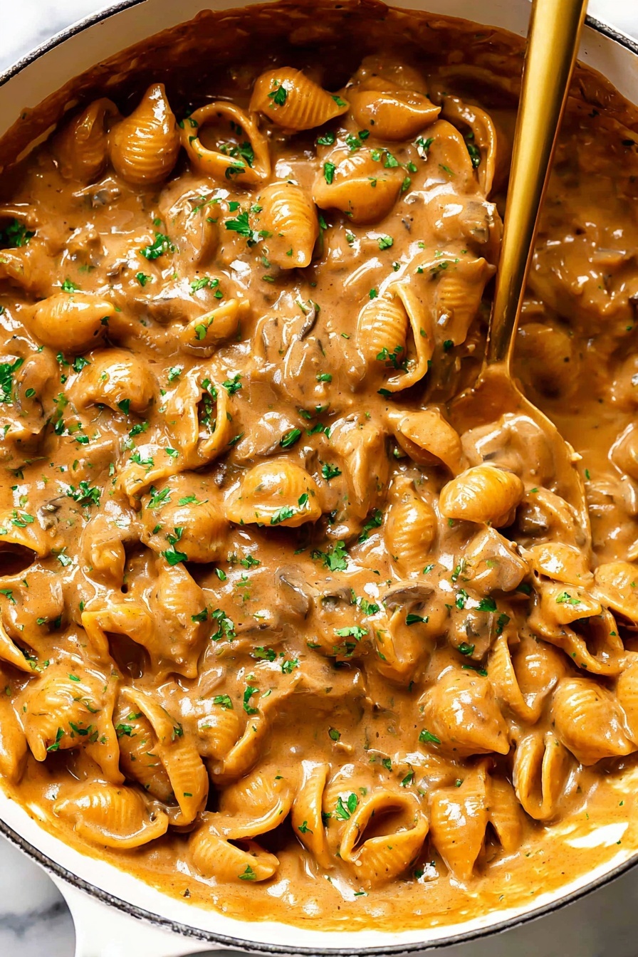 French Onion Pasta with Gruyere, French Onion Pasta recipe, caramelized onion pasta, cheesy pasta recipes, easy onion pasta dish - A close-up view of a white pan filled with a rich, creamy mushroom sauce that has a warm brown color and a thick texture. The sauce contains many small mushroom slices that create a bumpy surface, covered evenly in the glossy, smooth sauce. Bright green parsley bits are scattered on top, adding a touch of color contrast. A shiny golden spoon rests inside the pan, partially submerged in the sauce, showing the thick consistency as it drags some sauce along. The background features a white marbled surface. photo taken with an iphone --ar 2:3 --v 7