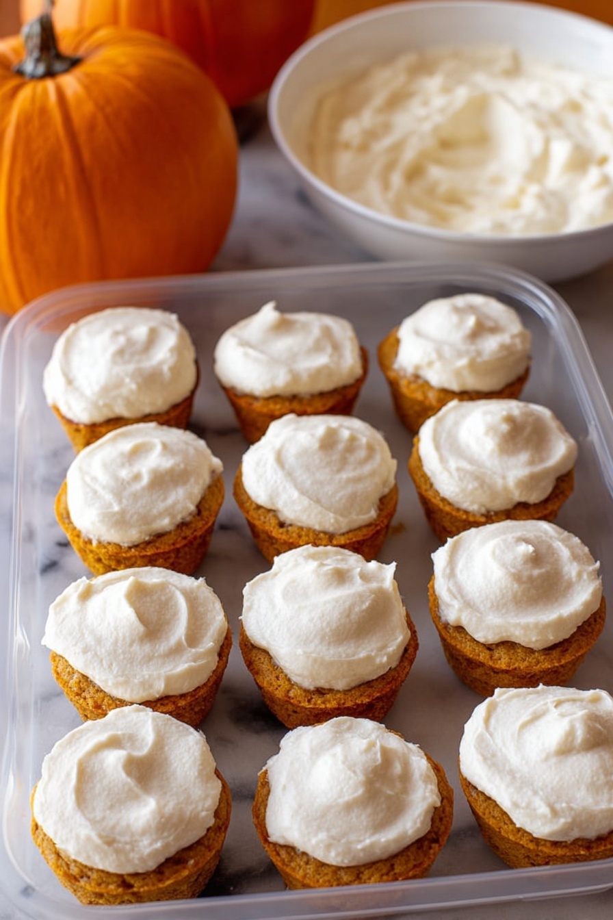 Flourless Pumpkin Muffins, Healthy Pumpkin Muffins, Grain-Free Pumpkin Muffins, Gluten-Free Pumpkin Muffins, Nourishing Pumpkin Snacks - The image shows a clear plastic container filled with twelve small cupcakes arranged in three rows of four. Each cupcake has two layers: a golden orange base with a slightly crumbly texture and a thick layer of white frosting spread smoothly on top. The frosting covers the entire top surface of each cupcake with a slightly swirled pattern. In the background, there is an orange pumpkin on the left and a white bowl filled with more white frosting on the right, all placed on a white marbled surface. photo taken with an iphone --ar 2:3 --v 7