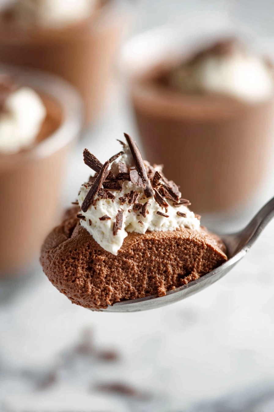 Decadent Chocolate Mousse, chocolate mousse recipe, rich chocolate dessert, silky mousse, easy chocolate mousse - The image shows a clear glass filled with a rich, smooth dark brown chocolate pudding. On top, there is a dollop of white whipped cream with delicate dark chocolate shavings scattered over it. The glass is placed on a white marbled surface, and in the background, there are blurred similar glasses filled with the same dessert. Photo taken with an iphone --ar 2:3 --v 7