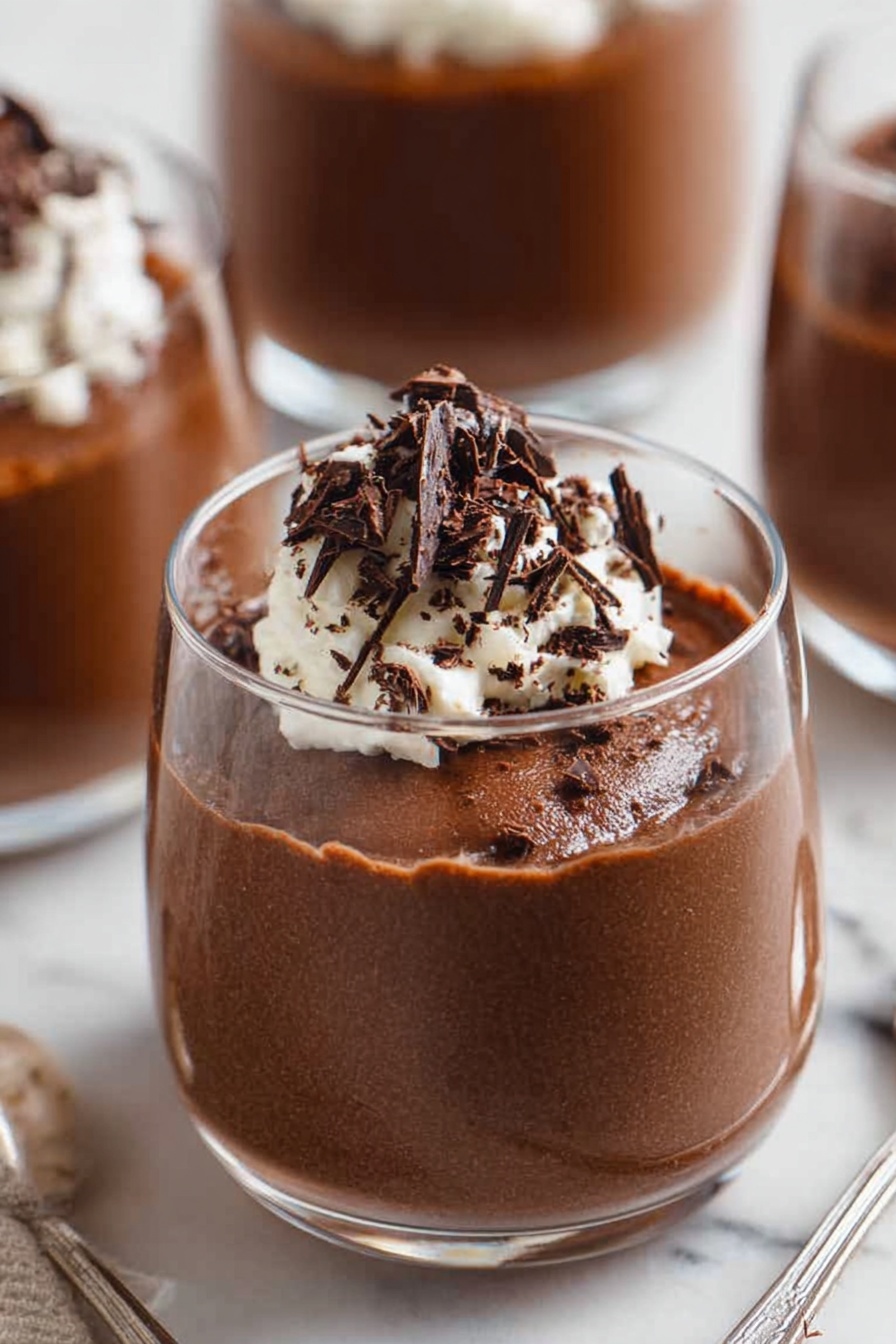 Decadent Chocolate Mousse, chocolate mousse recipe, rich chocolate dessert, silky mousse, easy chocolate mousse - A close-up of a spoon holding a scoop of light and airy chocolate mousse with a rough texture, topped with a small dollop of white whipped cream and thin, dark chocolate shavings scattered on top, with two blurry white bowls filled with the same mousse and cream in the background, all set on a white marbled surface. photo taken with an iphone --ar 2:3 --v 7