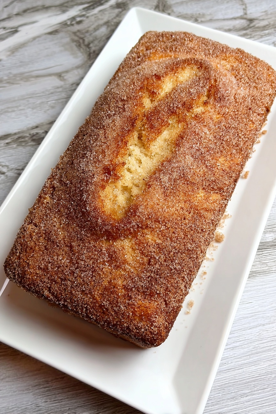 Cinnamon Donut Bread, cinnamon bread recipe, glazed bread with cinnamon, homemade cinnamon bread, easy cinnamon bread - A rectangular cake with a golden-brown top layer covered evenly with a fine, grainy light brown sugar coating sits on a white rectangular plate. The cake's top surface shows a gently cracked texture with soft ridges and swirls, revealing a tender and moist interior below the sugar crust. The plate rests on a white marbled texture surface. photo taken with an iphone --ar 2:3 --v 7