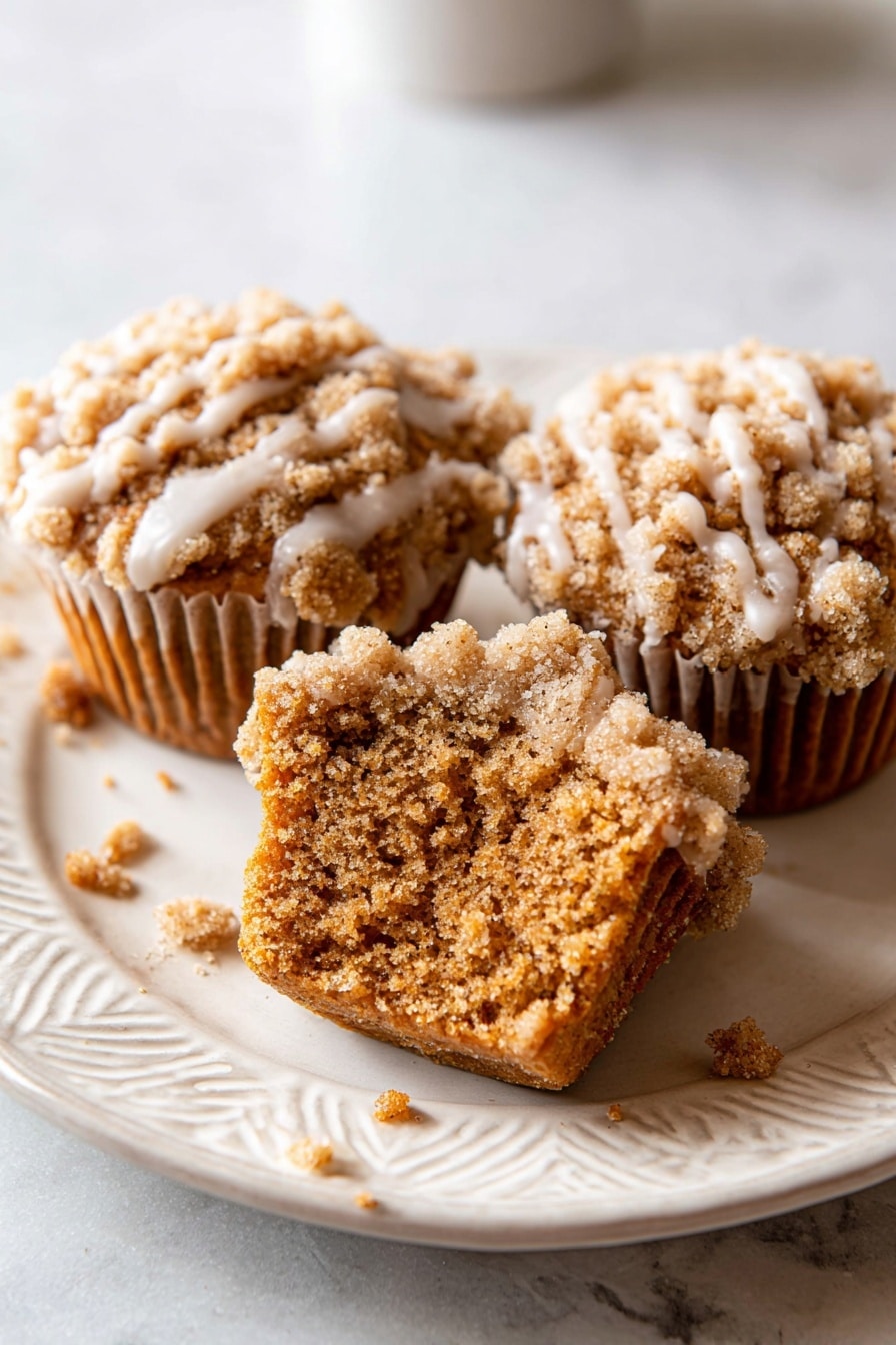 Pumpkin Crumb Cake Muffins, fall muffin recipes with pumpkin, easy pumpkin muffin recipe, moist pumpkin muffins with streusel topping, autumn breakfast treats - The image shows seven golden brown muffins arranged closely on a white plate. Each muffin has a crumbly, rough-textured top layer that is light brown and covered with a light cream-colored glaze that drips down the sides in thin streaks. The muffins are placed on a smooth white marbled surface underneath the plate. Some crumbs are scattered around the plate and surface. The lighting highlights the texture and glaze shine on the muffins. Photo taken with an iphone --ar 2:3 --v 7