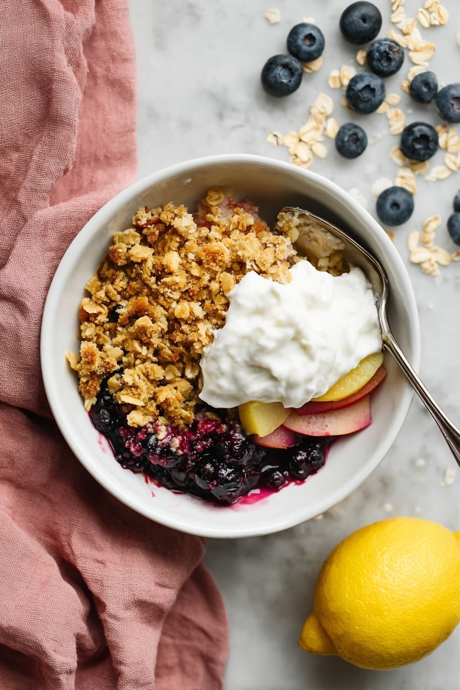 Apple and Blueberry Crumble, apple blueberry crumble dessert, easy fruit crumble recipe, fruit crisp with apples and blueberries, homemade berry crumble - A white bowl sits on a white marbled surface, filled with three main layers. The bottom layer contains dark blue and purple berries mixed with thin slices of light pink fruit. Above that is a thick, crumbly layer of golden brown oat crumble with hints of toasted oats on top. The final layer is a generous dollop of white whipped cream placed in the center of the crumble. A silver spoon is partly inside the bowl on the right side. Next to the bowl is a yellow lemon and scattered blueberries and oats, with a soft pink cloth to the left. photo taken with an iphone --ar 2:3 --v 7