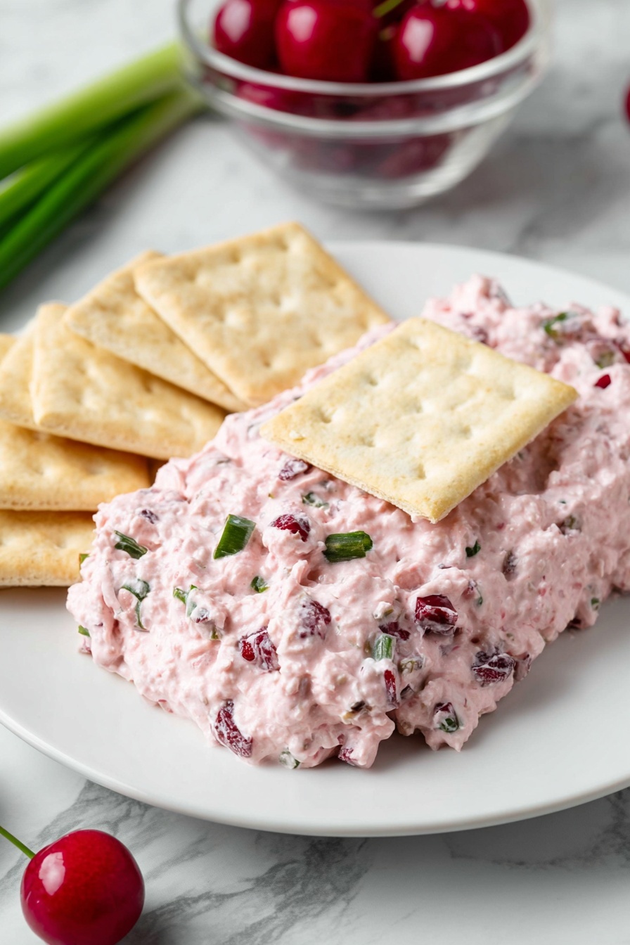 Cranberry Jalapeno Dip, Cranberry Jalapeno Dip recipe, spicy cranberry dip, holiday cranberry dip, easy party dip - On a white plate, there is a thick layer of pink creamy spread with small chunks of darker red and green mixed in. The spread is topped with a rectangular light beige cracker in the center. Around the spread, there are more light beige crackers placed on the white plate. The background shows a green onion on a white marbled surface and a clear bowl with red cherries. A green chili pepper is partially visible near the plate. photo taken with an iphone --ar 2:3 --v 7