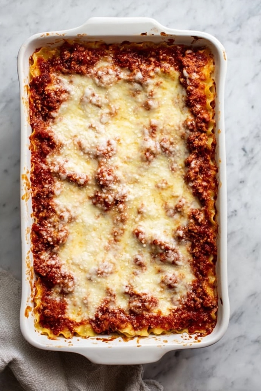 Vegan Lasagna Rolls, vegan lasagna rolls recipe, plant-based lasagna rolls, vegan stuffed pasta, vegan baked lasagna - A white rectangular ceramic baking dish filled with a baked lasagna, showing at least three layers: a bottom layer of pasta sheets, a middle layer of rich red tomato sauce mixed with browned ground meat, and a top layer covered evenly with melted, slightly browned cheese that looks soft and smooth. The edges of the dish have some browned sauce splashes. The dish sits on a surface with a white marbled texture, and a light-colored cloth is partially visible under the dish on the lower left side. Photo taken with an iphone --ar 2:3 --v 7