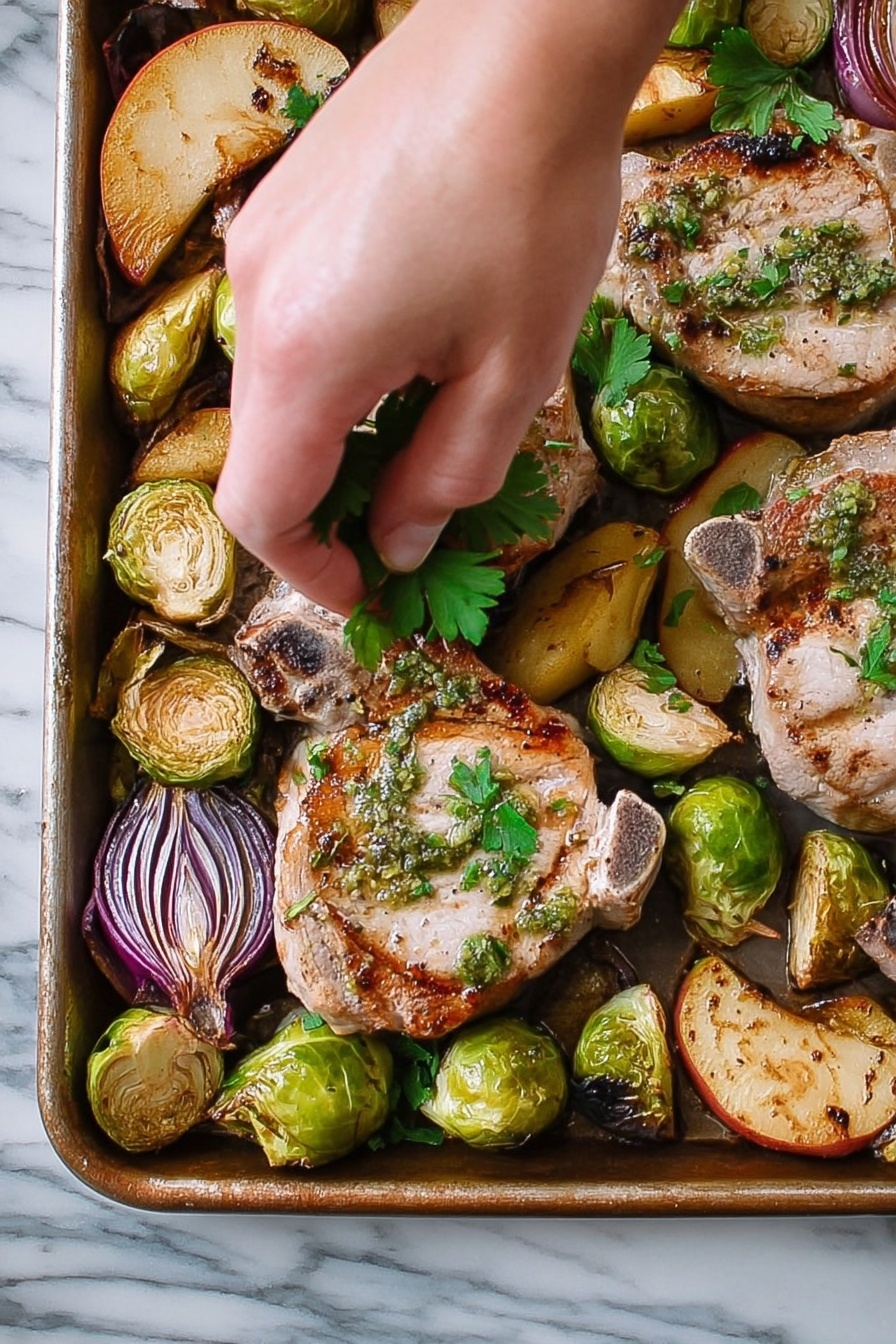 Sheet Pan Pork Chop Supper with Apple, Brussels Sprouts, and Sage, pork chop sheet pan dinner, easy pork chop recipes, cozy weeknight meals, apple and Brussels sprouts pork - A close-up view of a metal tray filled with cooked pork chops and roasted vegetables. There are three pork chops, each light brown with grill marks and green herb sauce on top, scattered across the tray. Around the pork chops, there are halved Brussels sprouts with a charred, darker green color, quartered pieces of red onions with purple layers, and golden brown apple slices. A woman's hand is gently placing fresh green parsley leaves over the pork chops. The tray sits on a white marbled surface. photo taken with an iphone --ar 2:3 --v 7