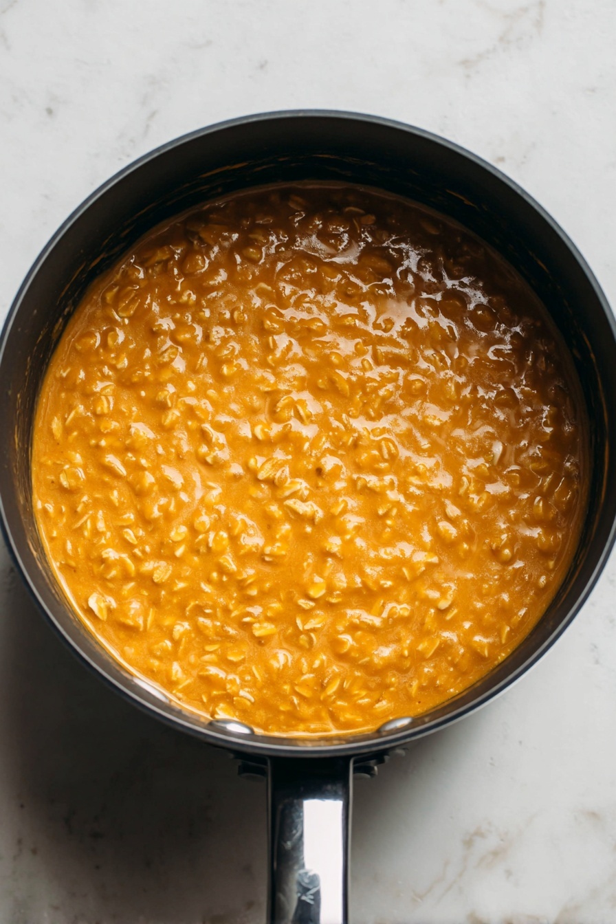 Pumpkin Oatmeal with Warm Spices and Nuts, fall breakfast ideas, cozy pumpkin oatmeal, healthy autumn breakfast, quick pumpkin oatmeal recipe - This image shows a close-up top view of a round black saucepan filled with a thick, creamy orange mixture with visible small oats throughout. The oats give the surface a bumpy texture, and the mixture looks smooth and warm. The saucepan is placed on a white marbled surface, and the shiny black handle is visible at the bottom right of the image. The lighting highlights the glossy shine of the mixture, making it look fresh and appetizing. photo taken with an iphone --ar 2:3 --v 7