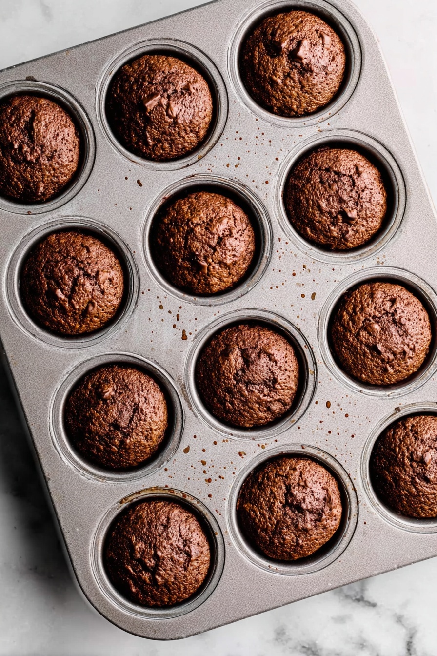 Sticky Toffee Pudding with Toffee Sauce, British dessert recipes, moist date pudding, easy toffee dessert, comforting British sweets - A metal muffin tray with 12 evenly spaced round muffins baked inside, each muffin a deep brown color with a rough, slightly cracked top texture showing some small chunks within. The muffins fit neatly in each circular slot of the tray, which shows small spots of light brown residue from baking. The tray is placed on a white marbled surface with soft natural lighting highlighting the muffins' golden and dark brown hues. photo taken with an iphone --ar 2:3 --v 7