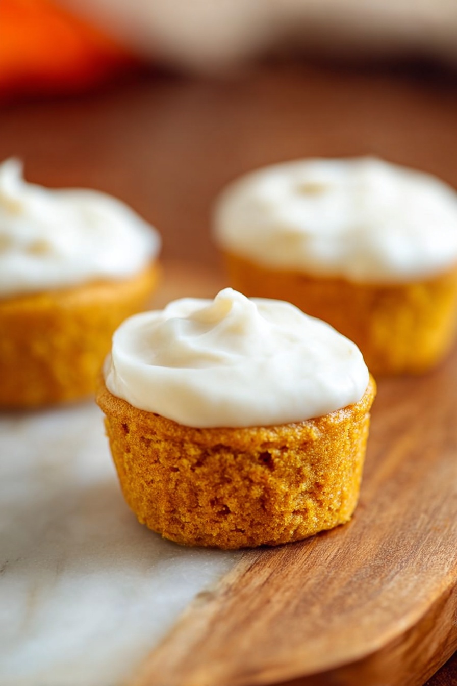 Flourless Pumpkin Muffins, Healthy Pumpkin Muffins, Grain-Free Pumpkin Muffins, Gluten-Free Pumpkin Muffins, Nourishing Pumpkin Snacks - Three small round cupcakes sit on a wooden surface with a white marbled texture. Each cupcake has one layer of golden orange cake with a soft crumb texture. On top of each cake, there is a smooth layer of white frosting spread evenly. The focus is on the closest cupcake in the front center, with the other two slightly blurred in the background. photo taken with an iphone --ar 2:3 --v 7