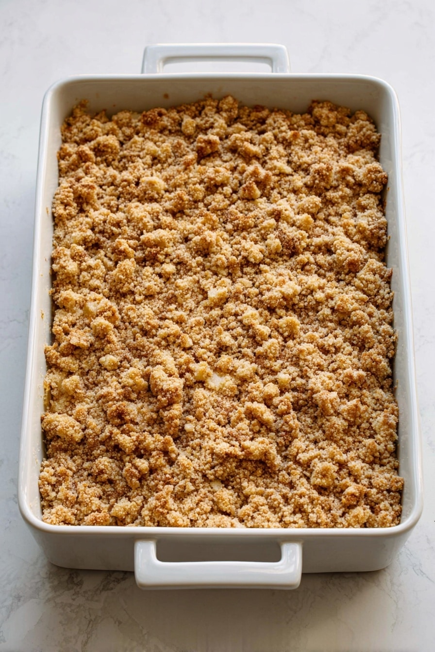 Caramel Apple Cheesecake Bars, caramel apple bars, apple cheesecake dessert, fall dessert recipes, easy cheesecake bars - A white rectangular baking dish with two handles is filled with a golden-brown crumbly topping that looks crunchy and uneven in texture, covering the entire surface. The topping has small clusters and a slightly rough look with hints of light and darker toasted shades. The dish is placed on a white marbled surface. photo taken with an iphone --ar 2:3 --v 7