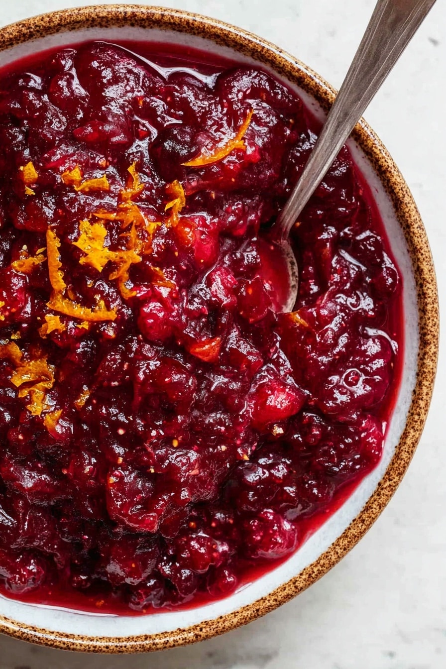 Easy Cranberry Sauce with Orange Juice, holiday cranberry sauce, simple cranberry sauce recipe, festive citrus cranberry sauce, homemade cranberry sauce - A close-up view of a thick, deep red cranberry sauce with a chunky texture, showing visible pieces of fruit mixed in. Small bits of bright orange zest are scattered on top, adding color contrast. The sauce fills a white bowl with brown speckled edges, and a silver spoon is placed inside the bowl, partly submerged in the sauce. The bowl sits on a white marbled surface. photo taken with an iphone --ar 2:3 --v 7