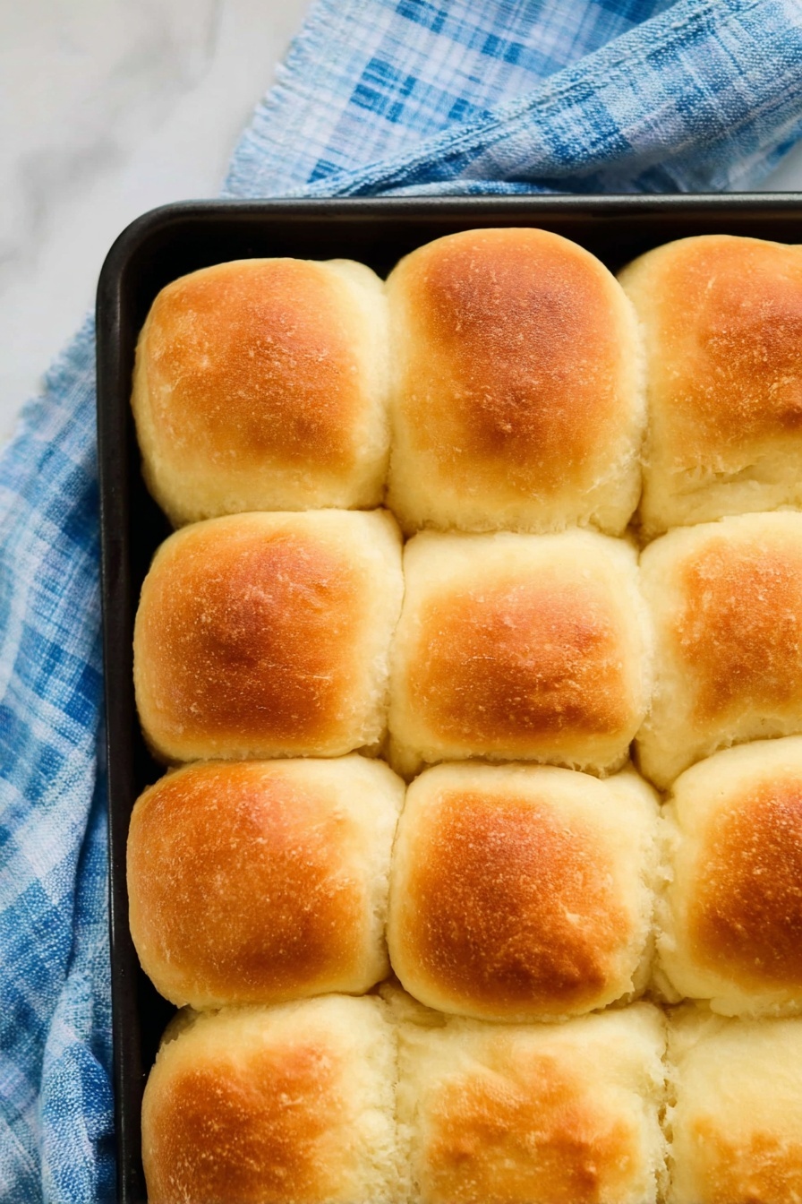 Vegan Dinner Rolls, vegan bread recipes, dairy-free dinner rolls, fluffy vegan rolls, plant-based dinner rolls - The image shows a close-up top view of sixteen golden-brown, fluffy bread rolls arranged in a 4 by 4 grid inside a black baking tray. Each roll has a slightly rounded top with a soft, smooth texture and a light crust that is golden on the upper part, fading to creamy white around the sides. The baking tray is placed on a blue and white checkered cloth on a white marbled surface. The rolls touch each other, showing their soft and puffy sides. photo taken with an iphone --ar 2:3 --v 7