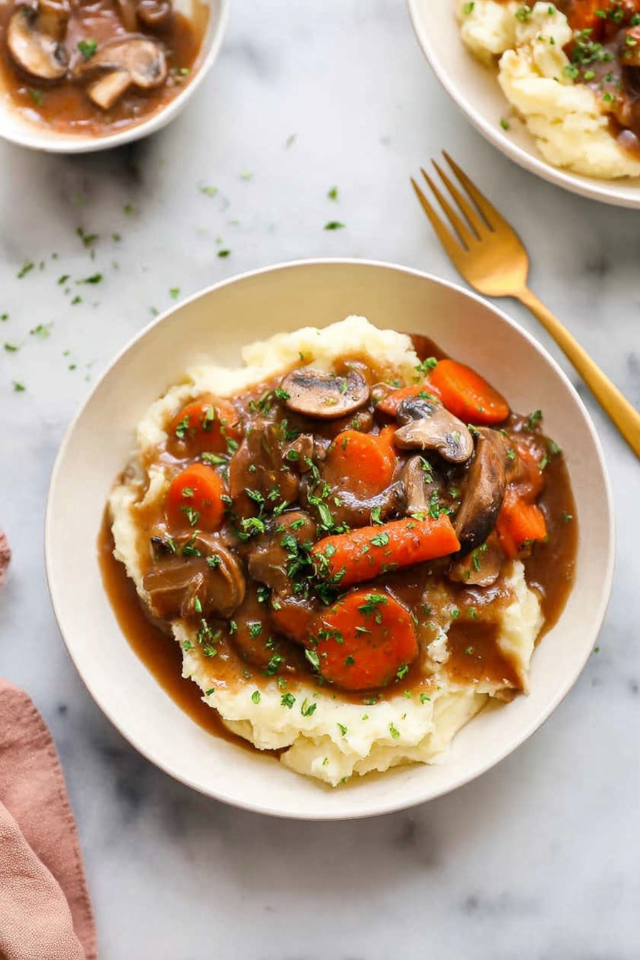 Mushroom Bourguignon, vegetarian mushroom bourguignon, easy mushroom stew, hearty mushroom dinner, flavorful mushroom recipe - A white bowl holds a dish with two main layers: the bottom layer is creamy mashed potatoes, soft and pale in color, covering the base of the bowl. On top is a rich brown gravy mixed with sliced mushrooms and large carrot chunks, with chopped green herbs sprinkled all over for color. A golden spoon and fork sit on the white marbled surface beside the bowl. In the background, another white bowl with the same dish is barely visible. The lighting is bright and natural, highlighting the textures and colors of the food photo taken with an iphone --ar 2:3 --v 7