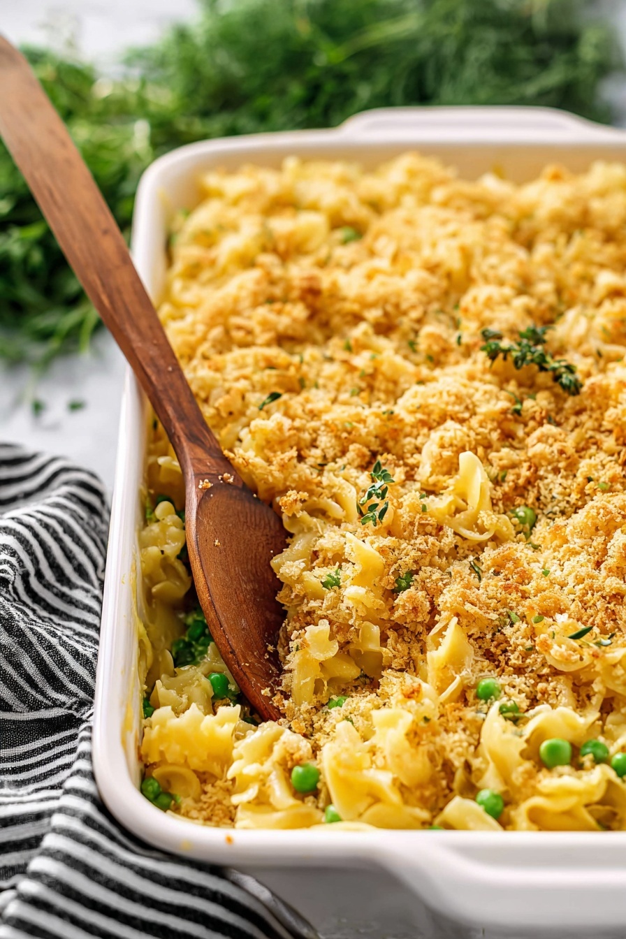 Creamy Chicken Casserole, Chicken Casserole, Easy Chicken Dinner, Comfort Food Recipes, Baked Chicken Dish - The image shows a white rectangular baking dish filled with a pasta casserole. The dish has three main layers: a bottom layer of yellow pasta, a middle layer with green peas mixed throughout, and a top layer covered with a golden brown crumbly topping that looks crispy. A wooden spoon stands inside the dish, scooping some pasta and crumbs. The background features a white marbled surface and some green herbs with a black and white striped cloth nearby. photo taken with an iphone --ar 2:3 --v 7