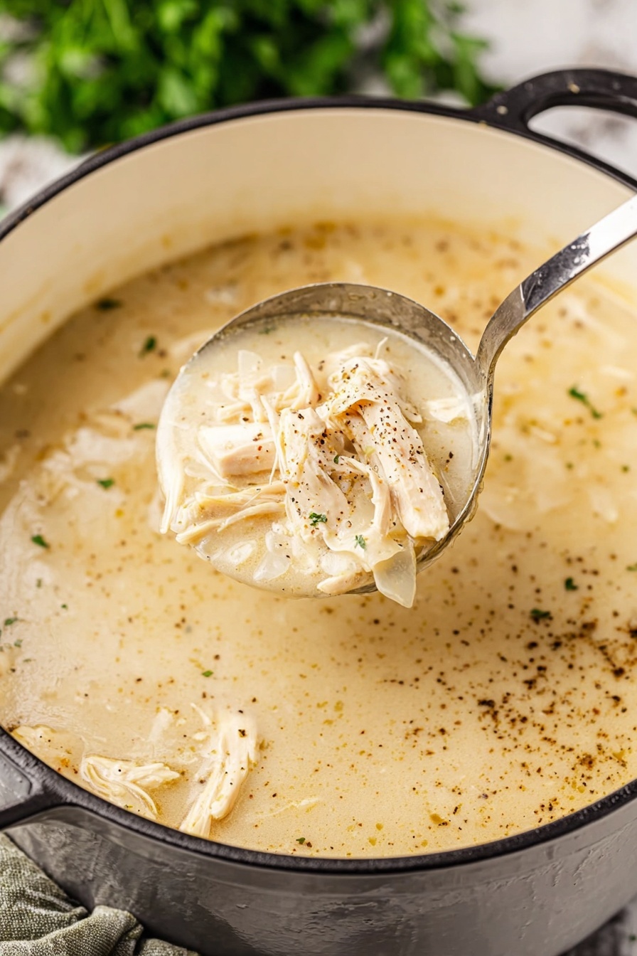 Southern Style Chicken and Dumplings, comforting chicken and dumplings recipe, homemade chicken and dumplings, Southern comfort food, easy chicken and dumplings - A close-up view of a creamy light beige soup with shredded light tan chicken pieces mixed in. The soup has a smooth, thick texture with visible specks of black pepper spread on the surface. A shiny metal ladle lifts a portion of the soup showing the creamy broth and chicken clearly. The soup pot is white inside with a black exterior handle visible, resting on a white marbled surface with green blurred herbs in the background. Photo taken with an iphone --ar 2:3 --v 7