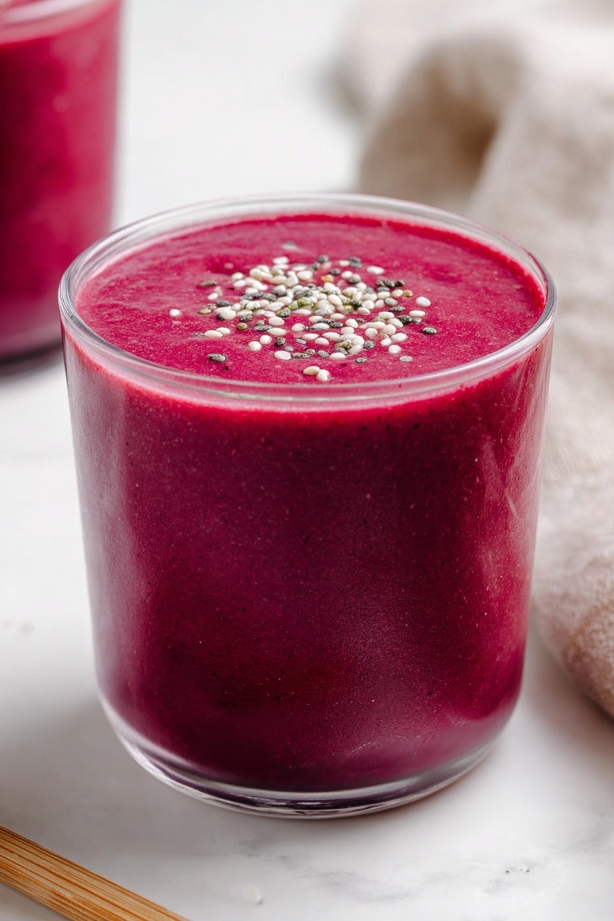 Banana Beet Smoothie, healthy beet smoothie, energizing fruit smoothies, nutritious smoothie recipes, beet and banana drink - A clear glass is filled with a thick, vibrant dark pink smoothie, almost reaching the top with a smooth and creamy texture. The smoothie surface is flat and topped with small white, black, and green seeds or grains sprinkled evenly across the center. The glass rests on a white marbled surface, with part of another glass filled with the same pink smoothie visible blurred in the background on the left. A light brown wooden stick lies horizontally near the base of the main glass on the left side, and a soft beige cloth is partly visible on the right side. Photo taken with an iphone --ar 2:3 --v 7