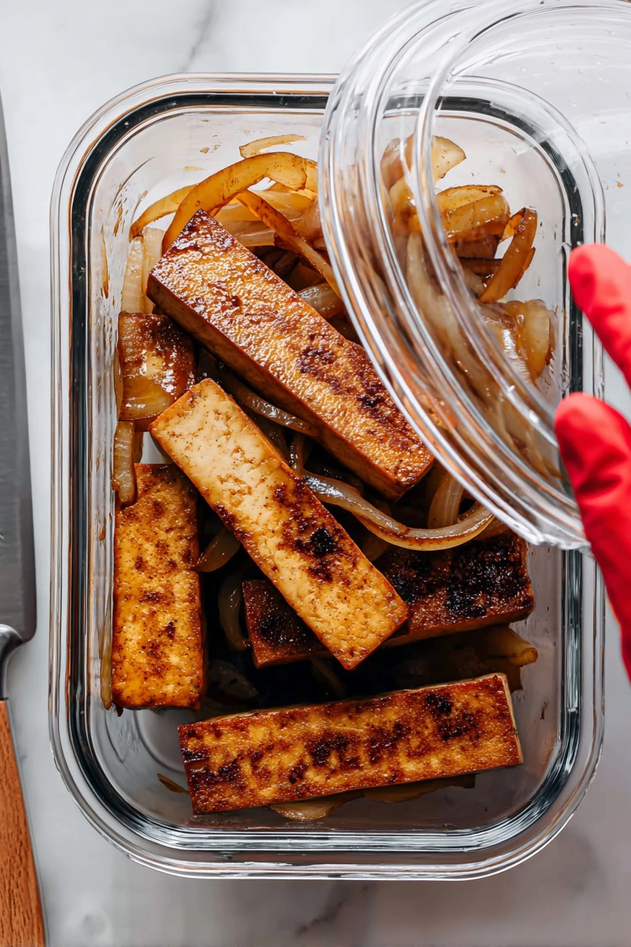 Sautéed Lime Tofu, Lime Tofu recipe, Crispy tofu with lime, Vegan lime tofu, Easy tofu stir-fry - The image shows a clear glass container holding several thick rectangular pieces of cooked tofu that are brown and have a slightly charred texture on some edges. Mixed among the tofu pieces are translucent sautéed onion slices that are light brown and curled. The container is placed on a white marbled surface, and a woman's hand with a red-gloved fingertip is lifting the clear lid. To the left, a silver knife rests on the surface partially visible. photo taken with an iphone --ar 2:3 --v 7