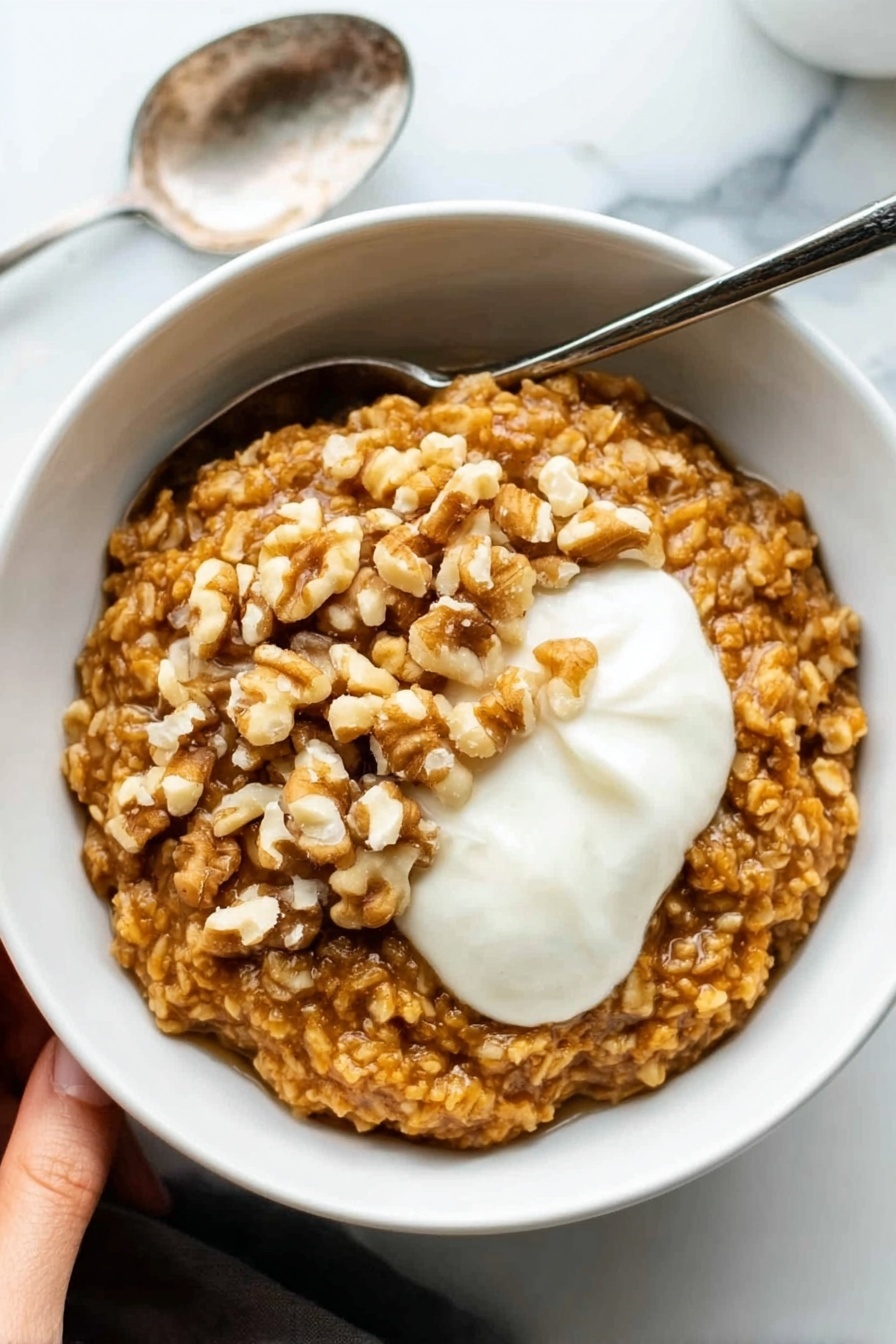 Pumpkin Oatmeal with Warm Spices and Nuts, fall breakfast ideas, cozy pumpkin oatmeal, healthy autumn breakfast, quick pumpkin oatmeal recipe - A white bowl filled with thick oatmeal that is golden brown in color and has a textured look from large oats. On top of the oatmeal, there is a layer of whole walnut pieces, adding a rough, uneven surface in light brown shades. Next to the walnuts, there is a dollop of smooth, white cream sitting on the right side of the bowl. The bowl is set on a white marbled surface, and a silver spoon rests next to it on the left. In the upper left corner, a woman's hand is visible holding the bowl. Photo taken with an iphone --ar 2:3 --v 7