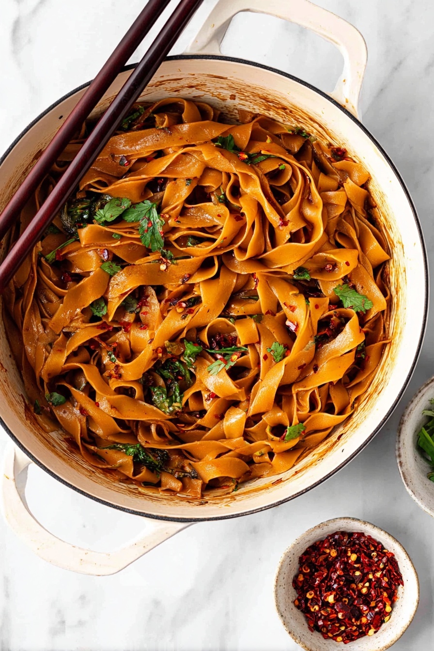 Spicy Chili Garlic Noodles, spicy Asian noodles, quick chili garlic pasta, spicy noodle stir-fry, flavorful chili garlic dish - A white bowl filled with wide flat noodles coated in a shiny reddish-brown sauce is shown from above. The noodles are mixed and curled inside the bowl with visible pieces of fresh green onions and chopped cilantro scattered on top. There are small white sesame seeds and dark chili flakes sprinkled over the noodles. A pair of dark wooden chopsticks rests on the edge of the bowl leaning inward. In the blurred background, two small white bowls hold red chili flakes and more chopped green onions. The surface is a white marbled texture. photo taken with an iphone --ar 2:3 --v 7