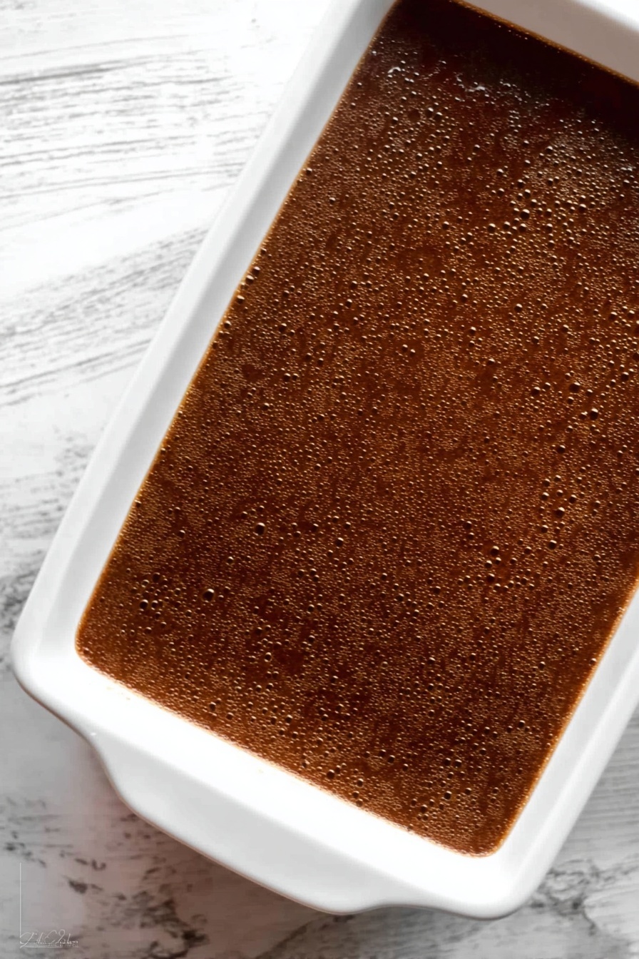 Gingerbread Cake with Butterscotch Sauce, gingerbread cake, spiced cake with butterscotch, cozy gingerbread dessert, moist gingerbread cake - The image shows a white rectangular baking dish filled with a smooth, dark brown dessert that has a few small bubbles on the surface, giving it a slightly textured look. The dish sits on a white marbled surface with visible veins. The dessert has a shiny, even top layer that reflects some light, indicating a moist texture beneath. Photo taken with an iphone --ar 2:3 --v 7