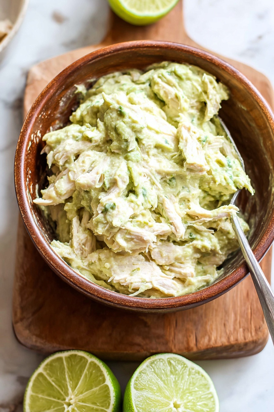 Healthy Greek Yogurt Avocado Chicken Salad, healthy chicken salad, avocado chicken recipe, Greek yogurt salad, keto friendly chicken salad - A brown bowl filled with a creamy, light green mixture that has a chunky texture with visible pieces of shredded white meat mixed inside. The bowl is placed on a wooden board, which rests on a white marbled surface. Behind the bowl, there are two halves of a lime, one facing up showing the green juicy interior, and the other turned to the side. A metal spoon is partially inserted into the mixture in the bowl. The scene is bright and fresh. photo taken with an iphone --ar 2:3 --v 7