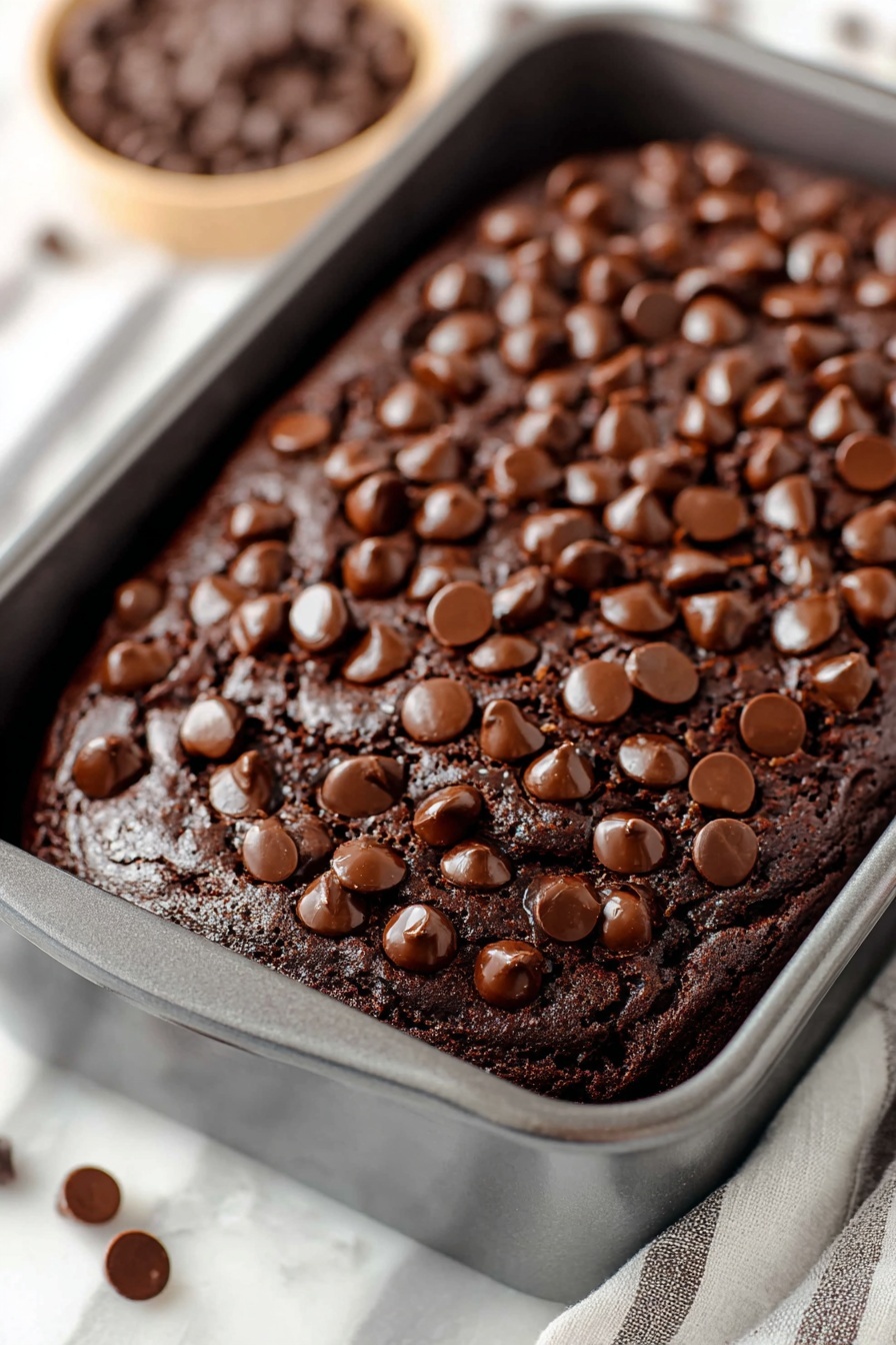 Chocolate Brownie Bread, brownie bread recipe, chocolate bread recipe, fudgy bread, easy brownie bread - A dark brown chocolate cake with a shiny cracked top fills a grey baking pan. The cake is covered with many round chocolate chips scattered all over the surface, adding texture and a shiny look. The edges of the cake are slightly raised, showing a soft, moist inside. The pan sits on a white marbled surface, with a bowl of chocolate chips blurred out in the background. Photo taken with an iphone --ar 2:3 --v 7