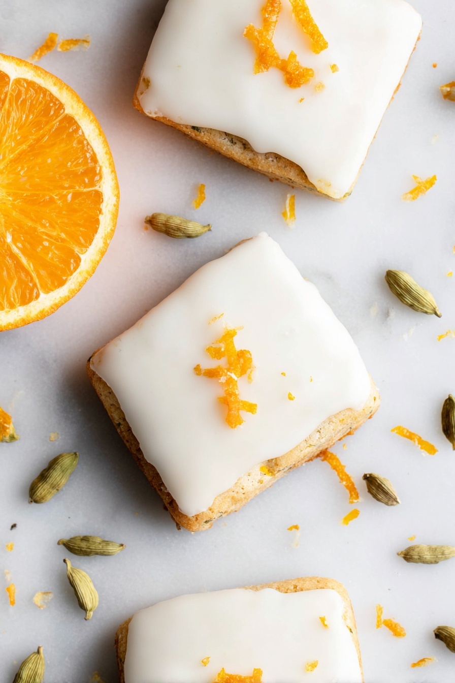Cardamom Shortbread Cookies with Orange Glaze, citrus shortbread cookies, flavored shortbread recipes, gourmet cookie recipes, elegant Christmas cookies - The image shows three square-shaped cookies with smooth white icing covering the top layer. Each cookie is slightly thick, with the base layer visible under the icing, showing a light golden-brown color and a slightly textured edge. Small pieces of orange zest are scattered on top of the icing, adding a bright orange color contrast. To the left side of the cookies, there is a fresh orange half with a vibrant, juicy orange color. Around the cookies on the white marbled surface, there are several light green cardamom pods and scattered bits of orange zest. The photo taken with an iphone --ar 2:3 --v 7