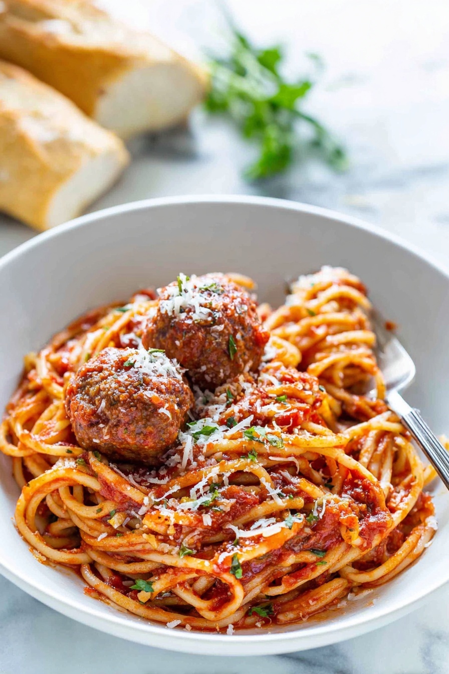 Classic Italian Spaghetti and Meatballs, easy spaghetti with meatballs, homemade Italian meatball recipe, authentic spaghetti and meatballs, best meatball pasta dish - A white bowl holds a serving of spaghetti noodles mixed with red tomato sauce, showing some herbs and seasoning. On top, there are three brown meatballs covered in sauce and sprinkled with white grated cheese. A silver fork rests inside the bowl on the right side. In the background, there is a blurred fresh green herb and some pieces of bread. The bowl sits on a white marbled surface. photo taken with an iphone --ar 2:3 --v 7