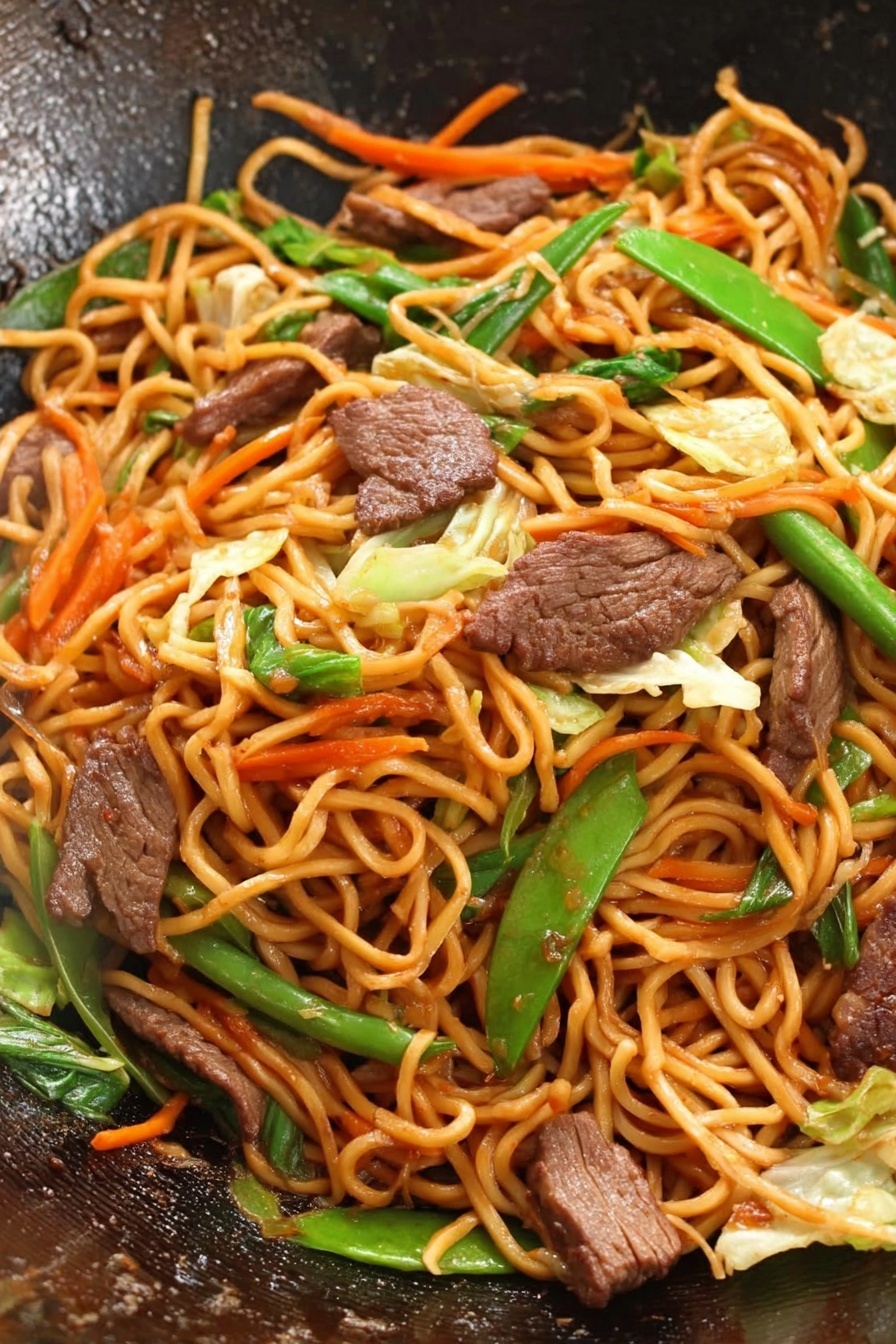 Beef Lo Mein Stir-Fry, quick beef stir-fry recipe, easy Chinese beef and noodle dish, flavorful veggie stir-fry, tender beef lo mein - This image shows a close-up of a wok filled with stir-fried noodles, meat slices, and mixed vegetables. The noodles are long and orange-brown, forming the main layer twisted together. Scattered throughout are medium-brown pieces of cooked meat, adding a rich texture. Bright green vegetables like sliced green onion and snap peas are placed evenly, with thin strips of orange carrot and light green cabbage mixed in, adding new colors and crunch. The surface inside the wok is dark and slightly shiny, highlighting the fresh, vibrant stir-fry. photo taken with an iphone --ar 2:3 --v 7
