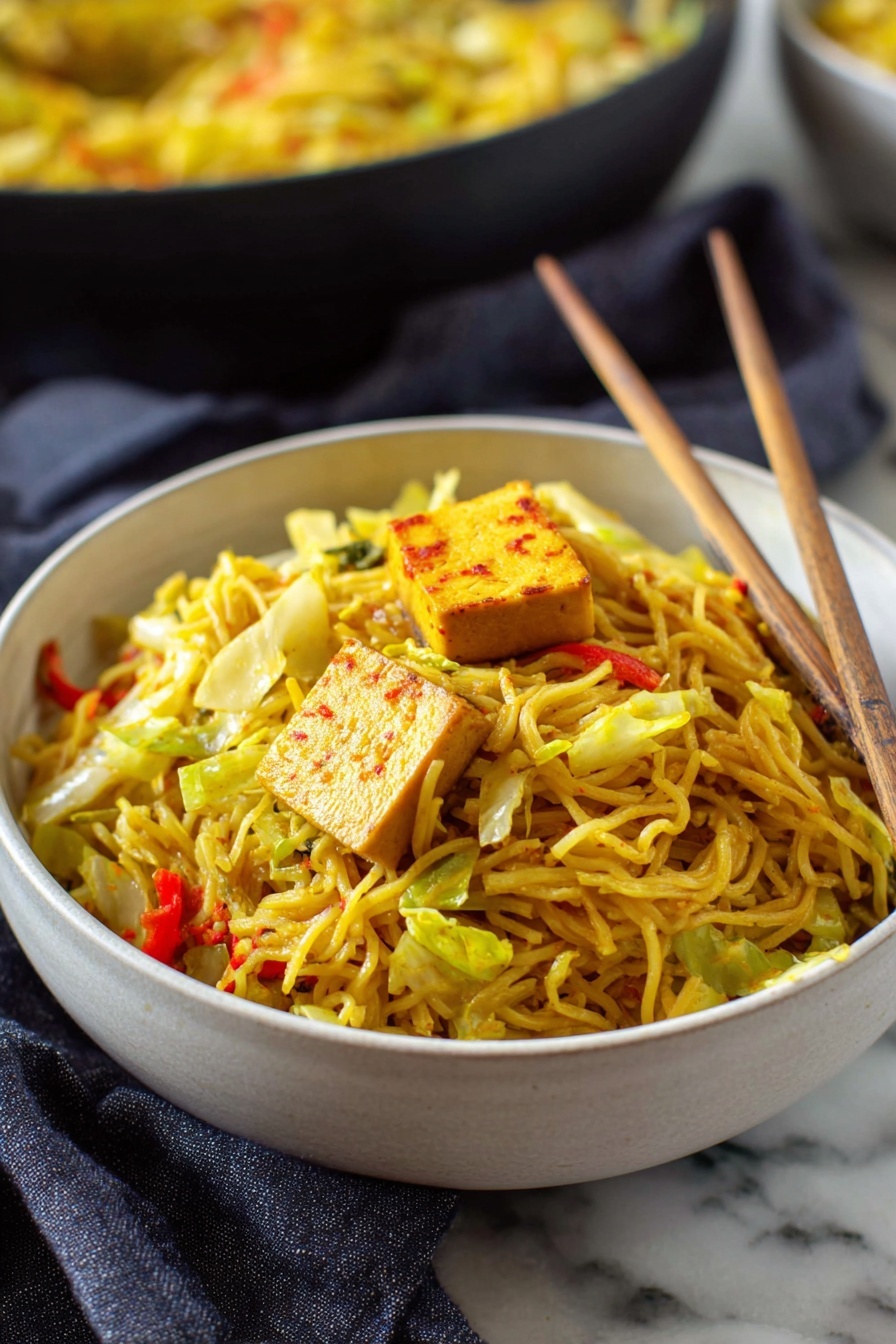 Easy Singapore Noodles, Singapore Noodles recipe, Asian noodle stir-fry, quick Singapore noodles, flavorful noodle dishes - A deep white bowl full of thin yellow noodles mixed with pieces of green cabbage and small bits of red bell pepper, topped with a square piece of light brown tofu with reddish edges. Light wooden chopsticks rest inside the bowl, leaning against the noodles on the right side. The bowl sits on a white marbled surface with a dark gray cloth nearby. In the background, there is another bowl filled with more noodles, blurred slightly. Photo taken with an iphone --ar 2:3 --v 7