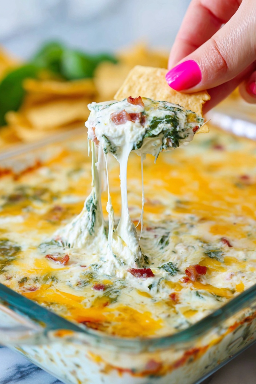 Bacon Spinach Artichoke Dip, spinach artichoke dip easy, cheesy bacon dip, party appetizer recipes, creamy bacon dip - A white square glass dish shows a baked dip with a top layer of melted golden-yellow cheese, lightly browned in spots. Under the cheese, thick creamy white sauce mixed with green spinach leaves and small bits of reddish-brown bacon is visible. A woman's hand with pink painted nails holds a triangular beige chip pulling up a string of stretchy melted cheese from the dish, showing the layers clearly. The background is blurry with hints of green and more chips. The whole scene is set on a white marbled surface. photo taken with an iphone --ar 2:3 --v 7