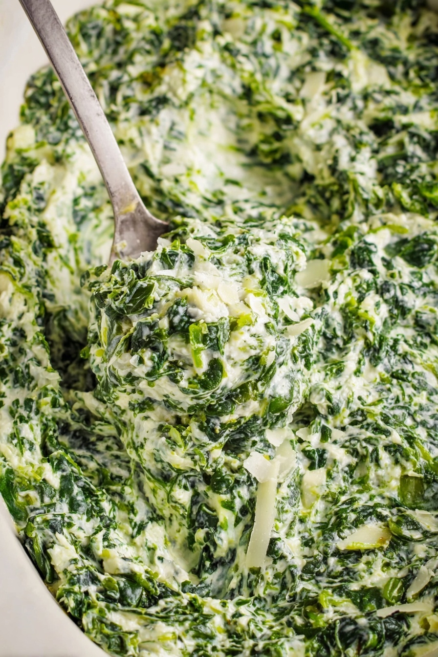 Creamed Spinach, Creamed Spinach Healthy, Creamed Spinach Easy, Creamed Spinach Side Dish, Creamed Spinach Cheesy - A close-up view of creamy spinach dip with visible layers of finely chopped green spinach mixed with white creamy sauce and small pieces of onion. The texture looks thick and rich with bits of grated white cheese sprinkled on top. A metal spoon scoops up a generous portion of the dip, showing the mix's dense, slightly chunky consistency. The dish sits on a white marbled surface that contrasts with the deep green and white colors of the dip. Photo taken with an iphone --ar 2:3 --v 7