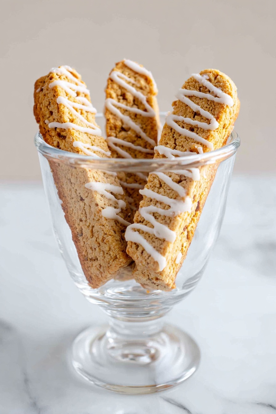 Cinnamon Vanilla Biscotti, biscotti recipe, homemade biscotti, coffee pairing cookies, crunchy vanilla biscotti - The image shows a clear glass cup placed on a white marbled surface. Inside the cup, there are three crispy biscotti standing upright, each with a rough light brown texture. White icing is drizzled in thin stripes across each biscotti, adding contrast to their warm golden color. The background is plain and softly lit, emphasizing the cup and its contents. photo taken with an iphone --ar 2:3 --v 7