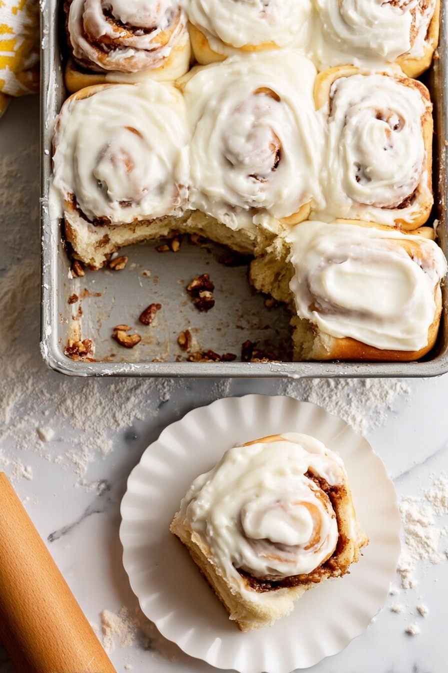 Soft Apple Cinnamon Rolls in Brioche Dough, apple cinnamon roll recipe, brioche cinnamon rolls, easy apple cinnamon rolls, homemade cinnamon rolls with apples - A metal baking dish holds six soft cinnamon rolls, with one removed showing the bottom layer coated in a sticky cinnamon filling and bits of nuts. Each roll has a creamy white icing thickly spread on top, swirled smoothly over the soft golden brown dough, with visible cinnamon spirals peeking through under the icing. One cinnamon roll sits on a small white scalloped plate in the lower right corner, showing its fluffy layered inside. The background is a white marbled surface lightly dusted with flour, with a wooden rolling pin nearby. photo taken with an iphone --ar 2:3 --v 7